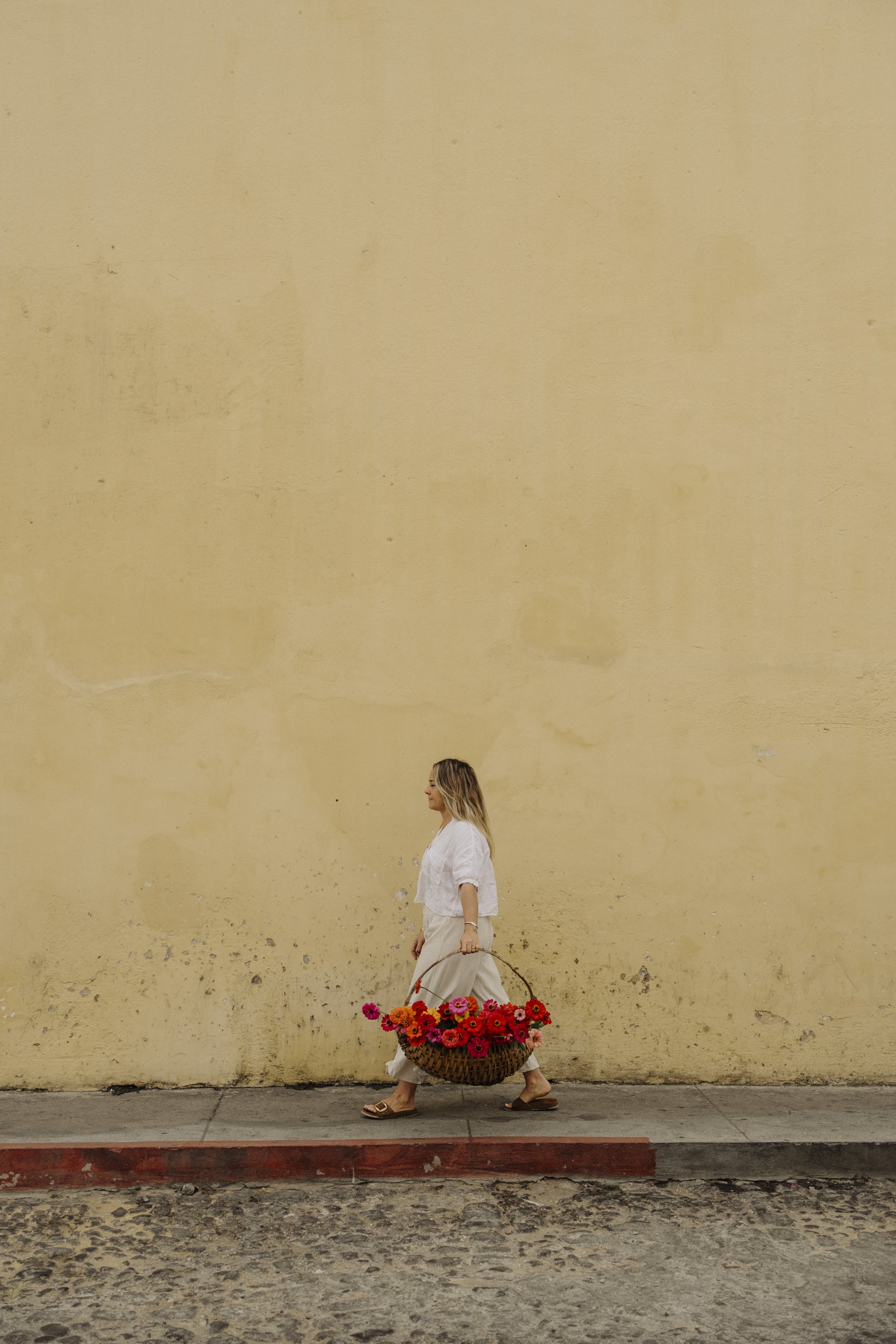 A woman with blonde hair walking along a sidewalk carrying a large basket filled with pink, orange, and red flowers. She wears a white shirt, beige pants, and brown sandals. The background is a plain beige wall.