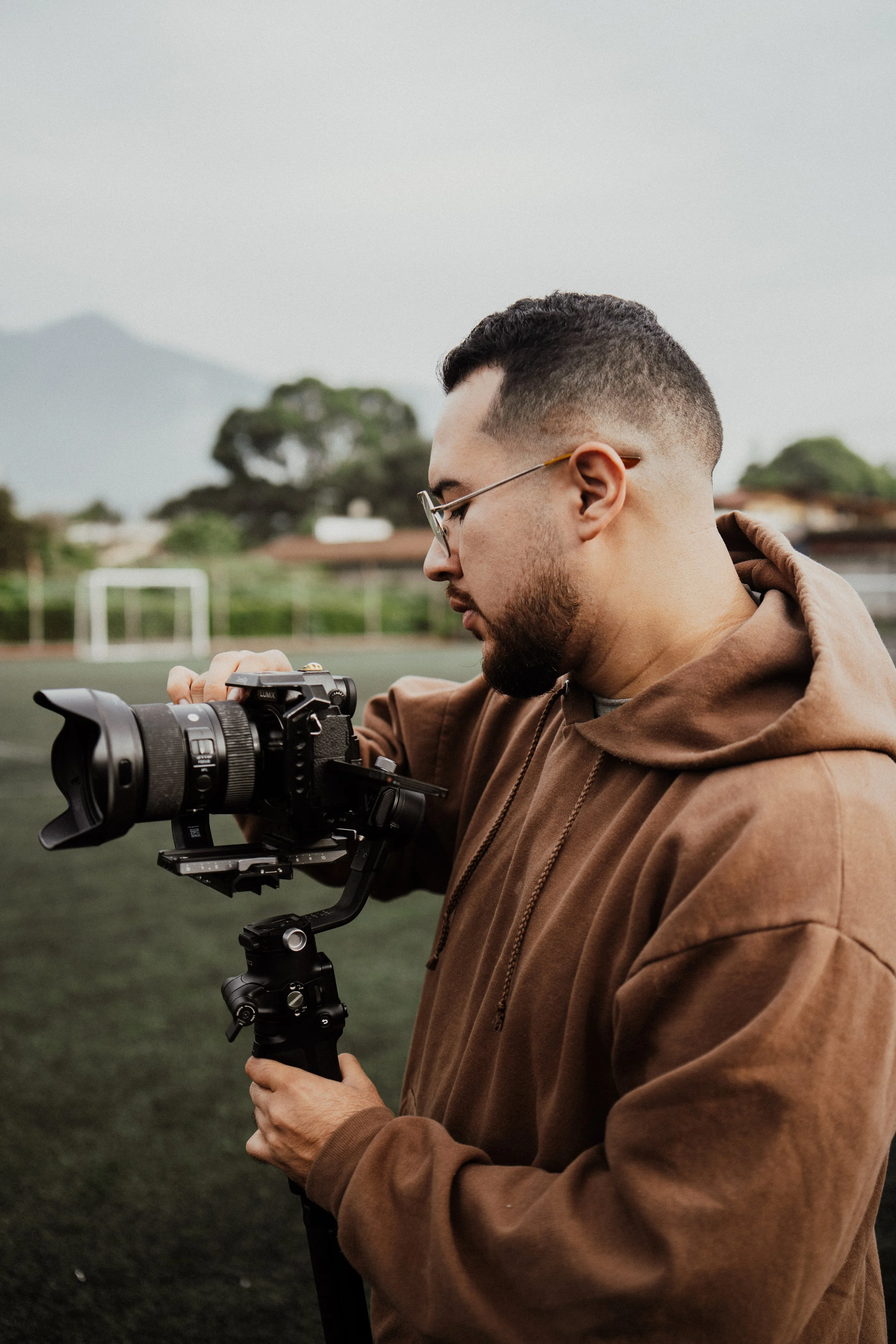 A man with glasses, a beard, and short dark hair wearing a brown hoodie is holding a camera mounted on a stabilizer, focusing on it during daytime on a sports field with goalposts and trees in the background.
