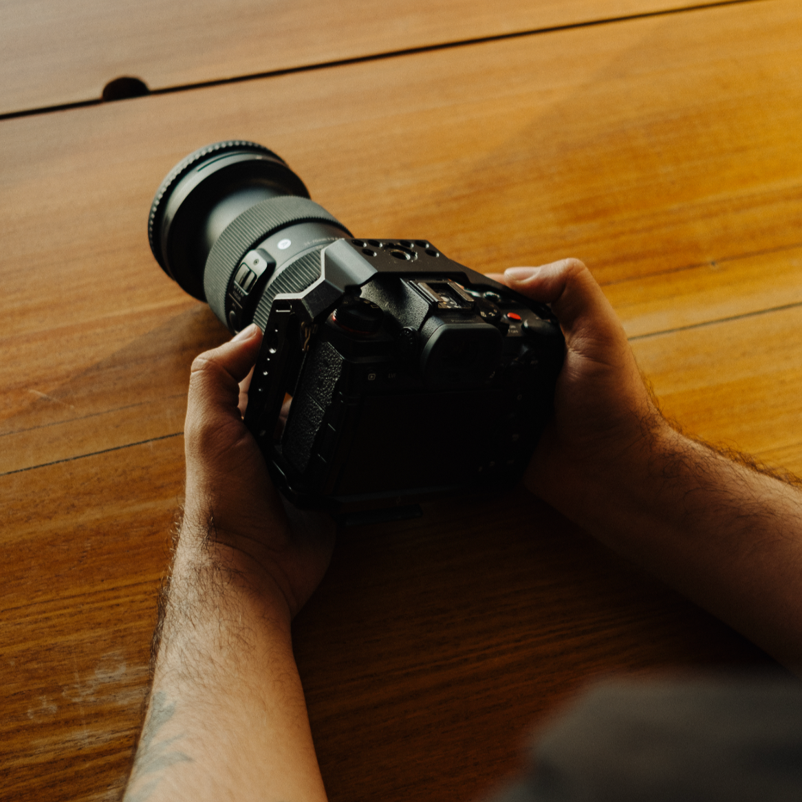 Close-up of a person holding a professional digital camera with a telephoto lens, resting on a wooden surface.