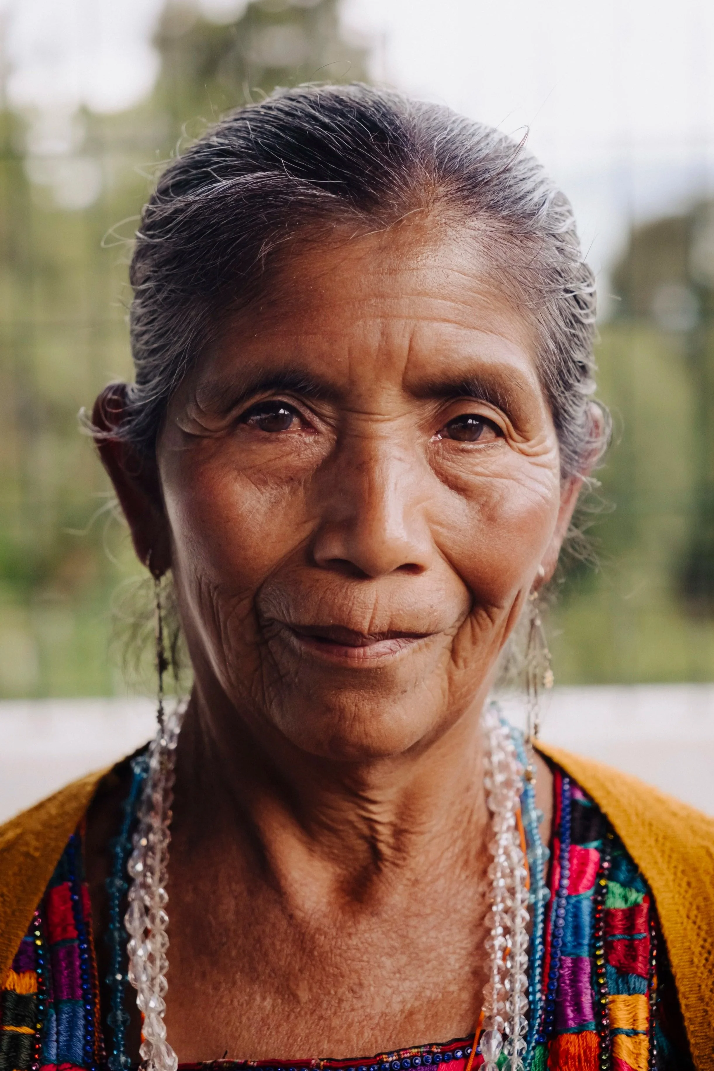 Close-up of an elderly woman with gray hair, wearing colorful traditional clothing, earrings, and necklaces, smiling gently.