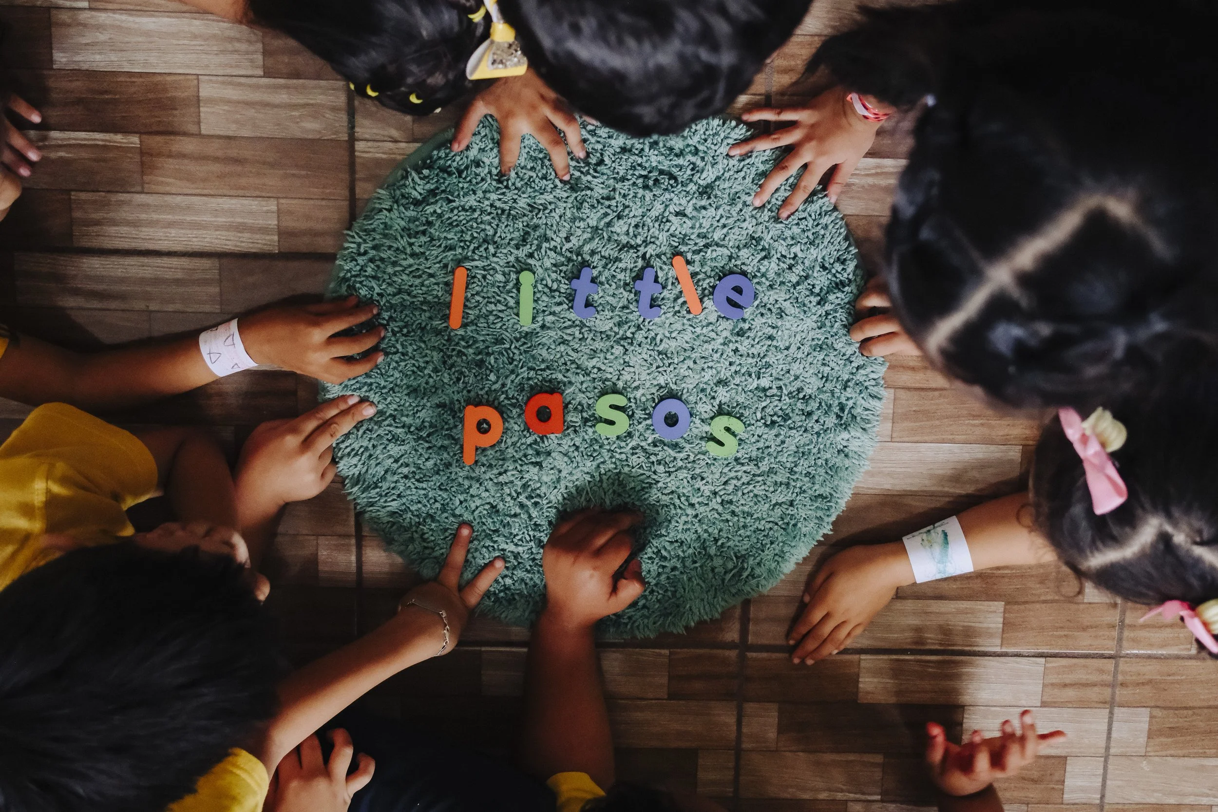 Children gather around a fuzzy green blanket with colorful letters spelling 'little pass' on it, on a wooden floor.