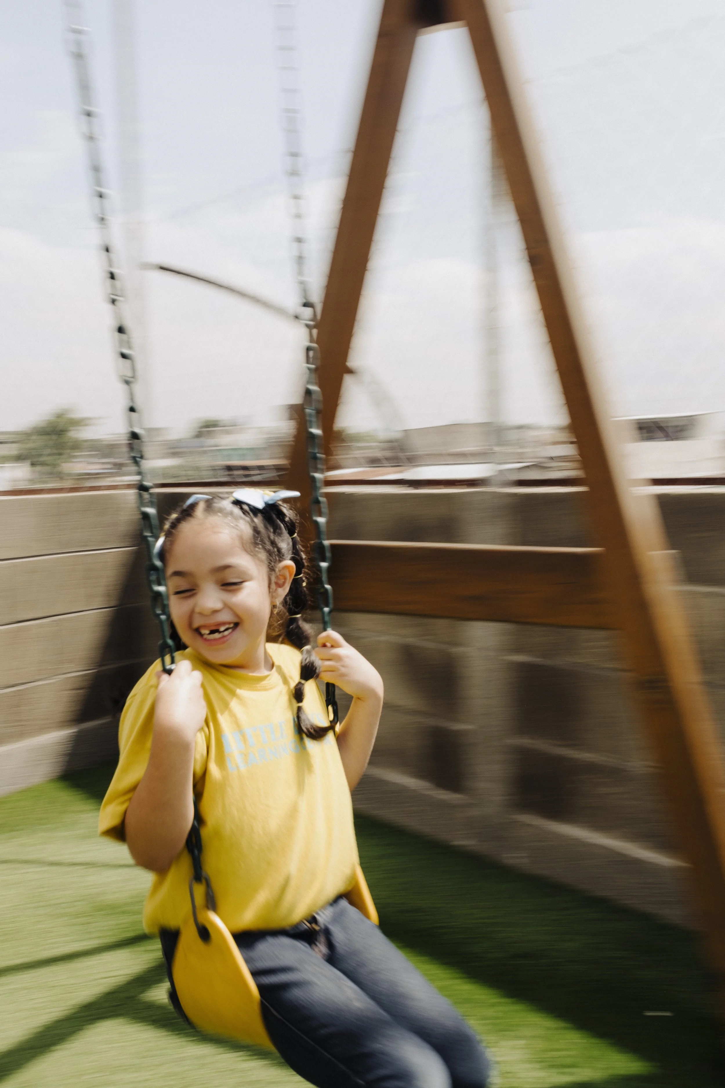A young girl is smiling and enjoying her swing ride at a playground. She has curly hair tied with a bow, and she is wearing a yellow T-shirt and dark pants. The swing is made of black chains and a yellow seat. The background shows a wooden fence and blurred structures.