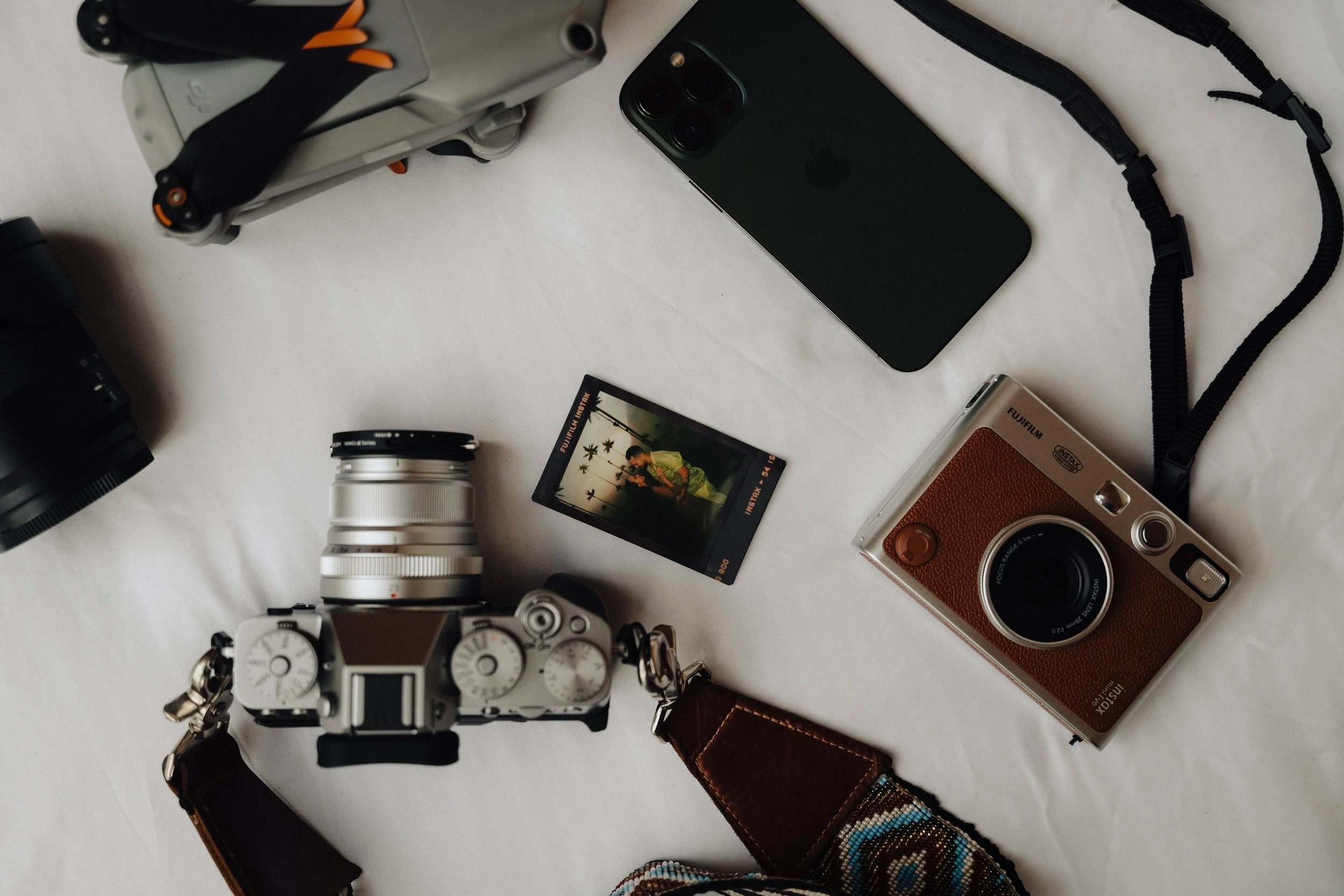 A flat lay of various photography and tech gadgets on a white surface, including a camera, a smartphone, a camera lens, a wristwatch, a film camera, a camera strap, and some camera accessories.