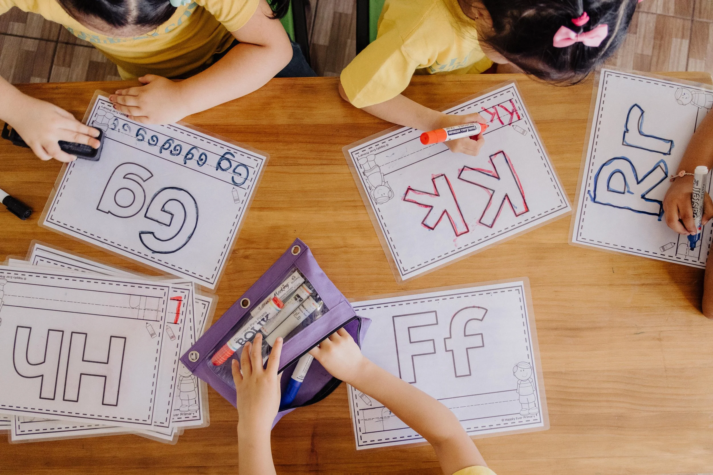 Children coloring and writing on worksheets with large printed letters, seated at a wooden table.