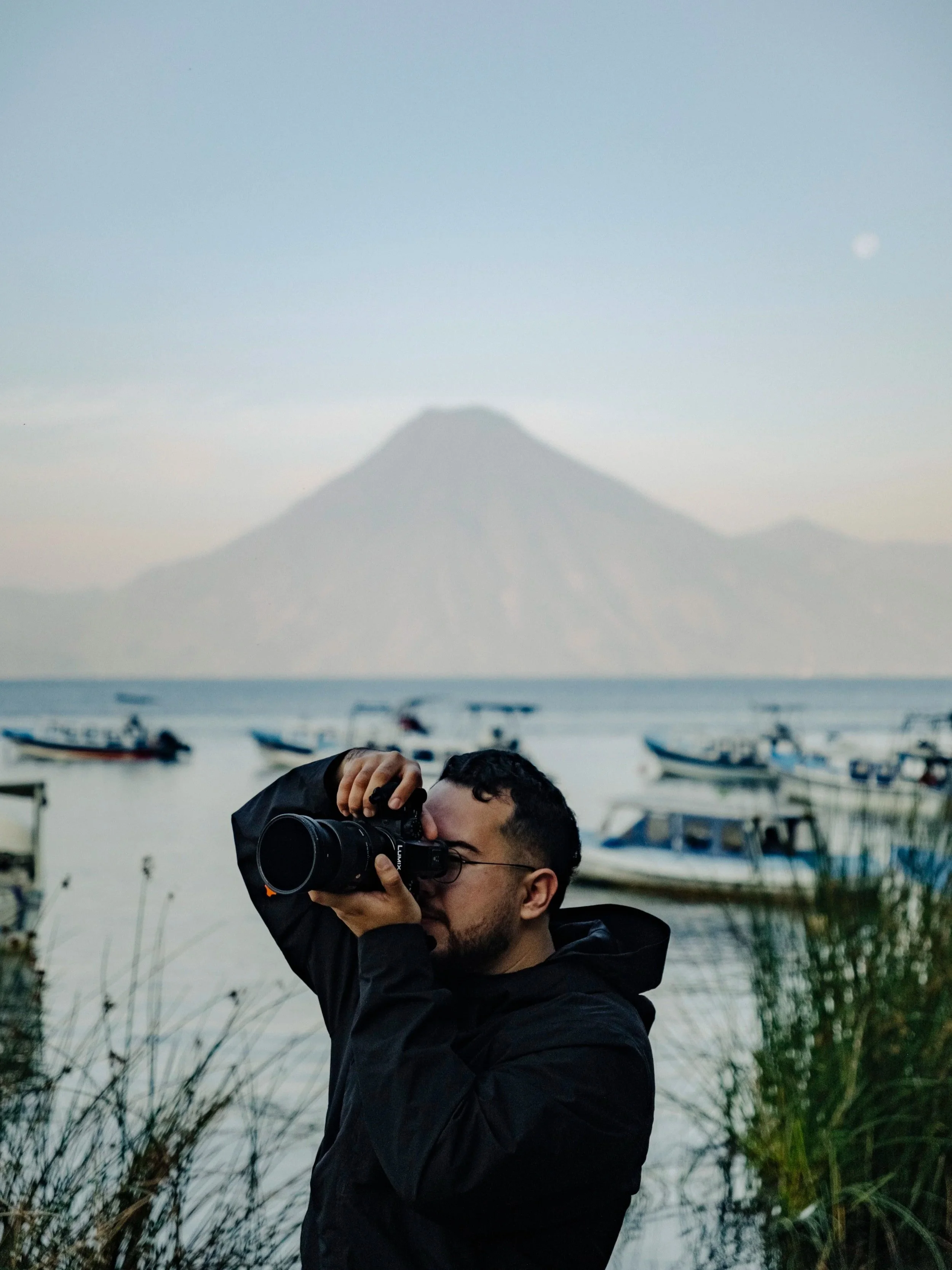 A person taking a photograph outdoors near water with boats docked and a mountain in the background during dusk.