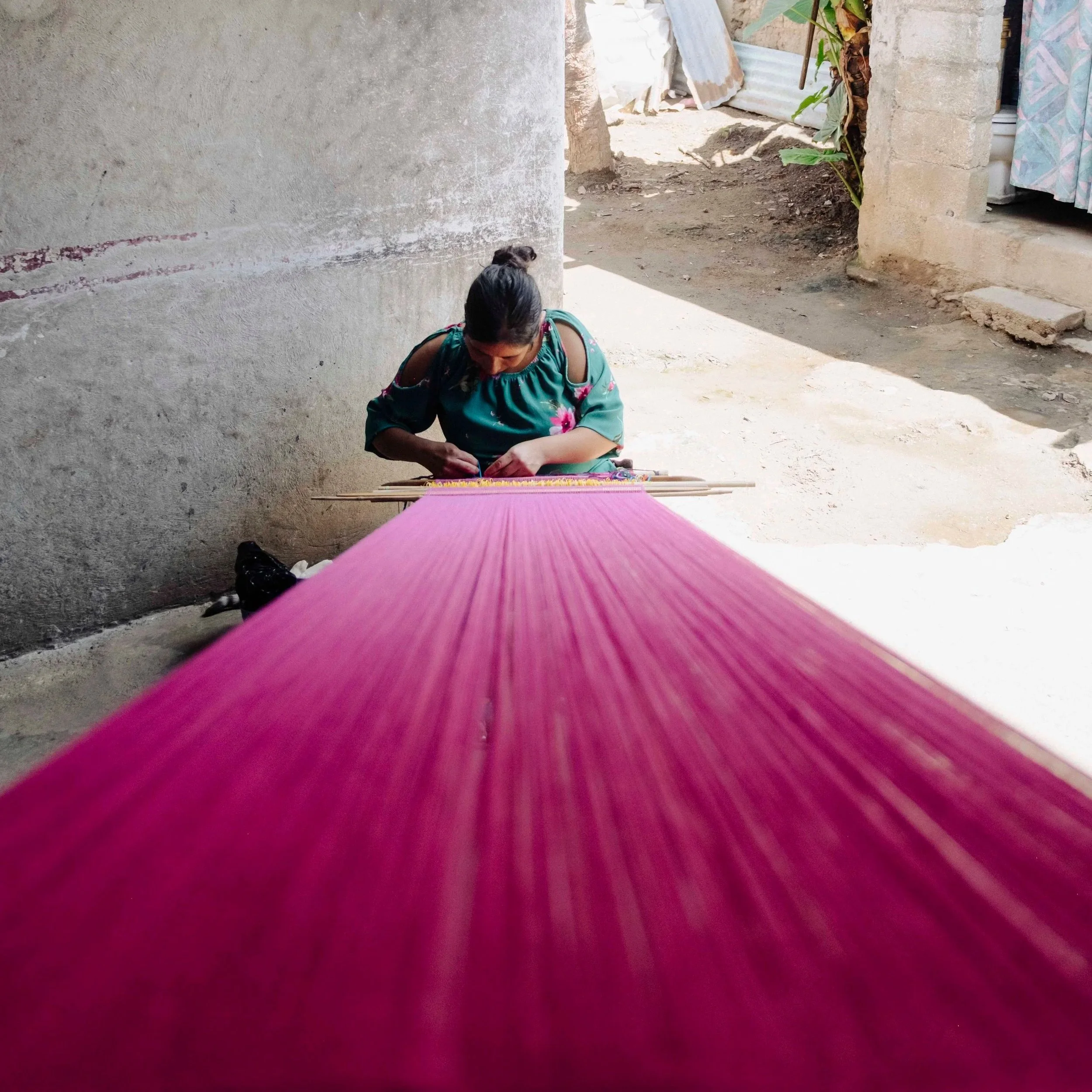 A woman wearing a green and pink floral dress sitting outdoors near a concrete wall, working on a colorful woven textile on a pink loom.