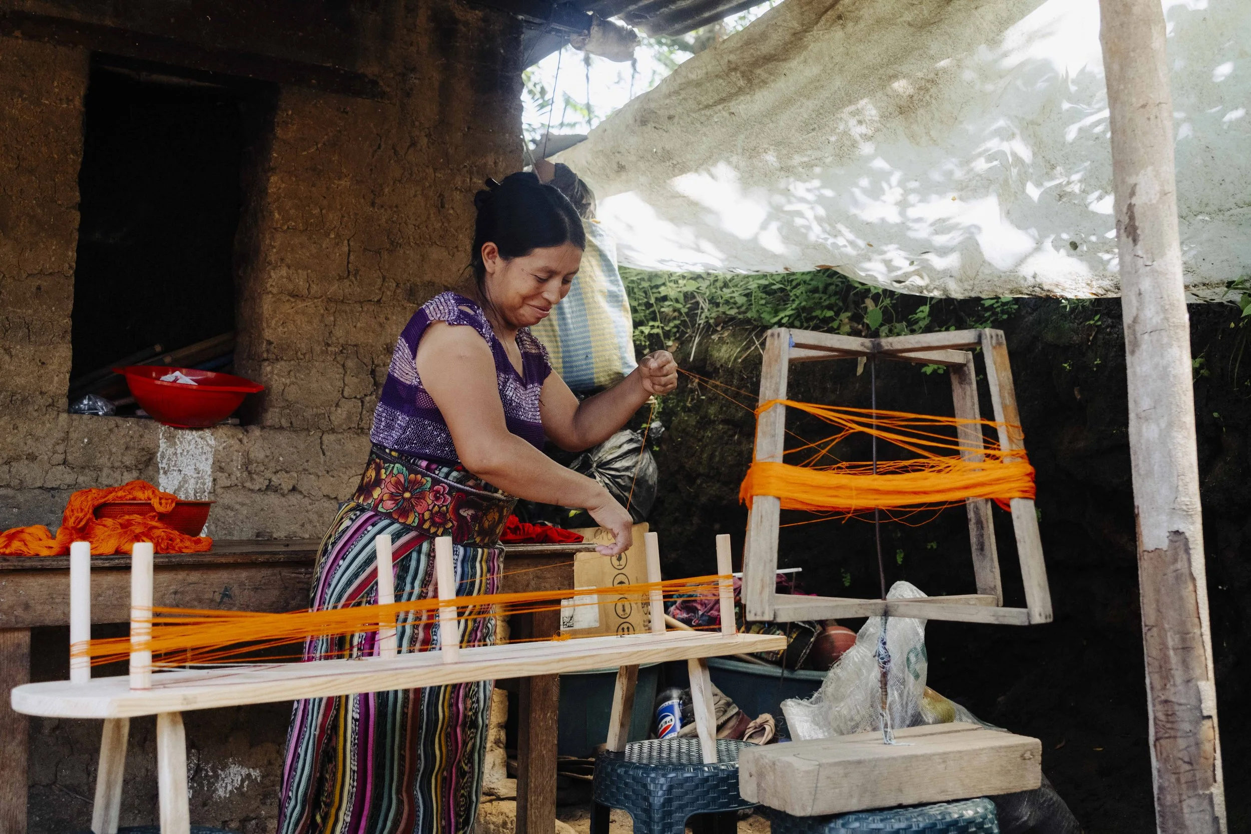 A woman with dark hair, wearing a colorful striped skirt and a purple top, is weaving orange thread on a wooden loom outdoors near a rustic mud wall. There are various colorful textiles and baskets around her, with a large white fabric hanging overhead and some greenery in the background.