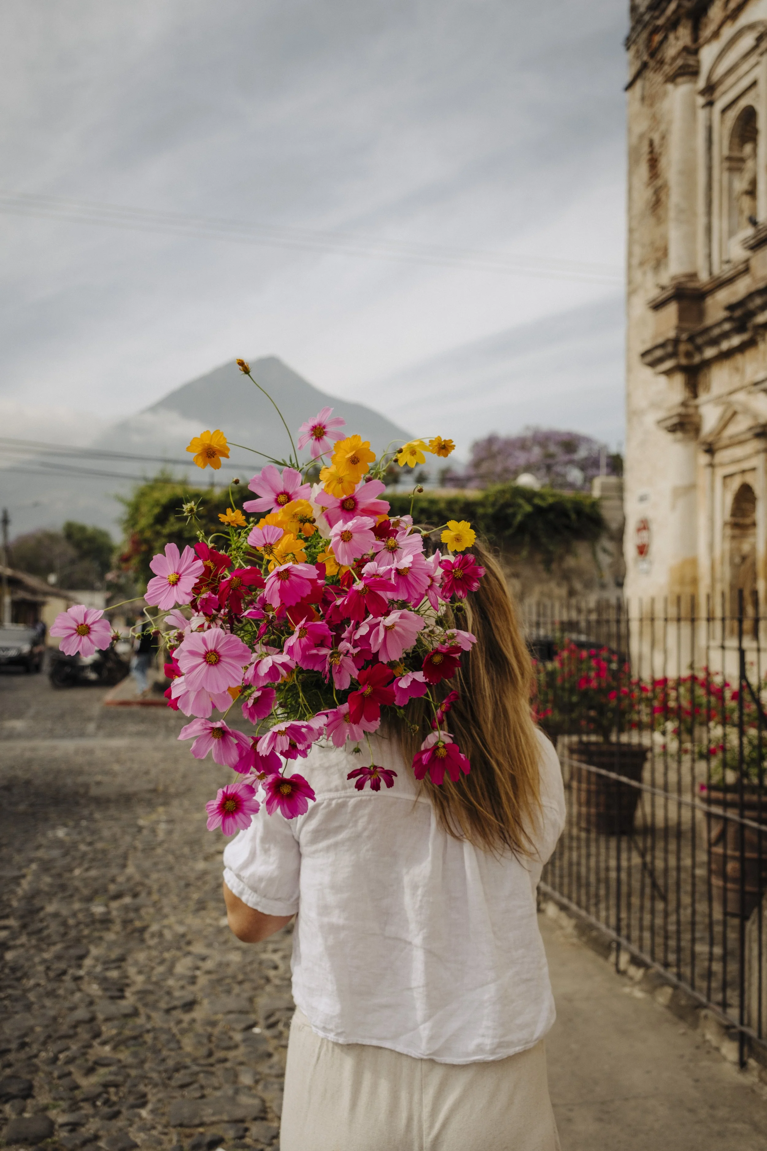 Person holding a large bouquet of pink, yellow, and orange flowers covering their face, standing outdoors near a historic stone building with a mountain in the background.