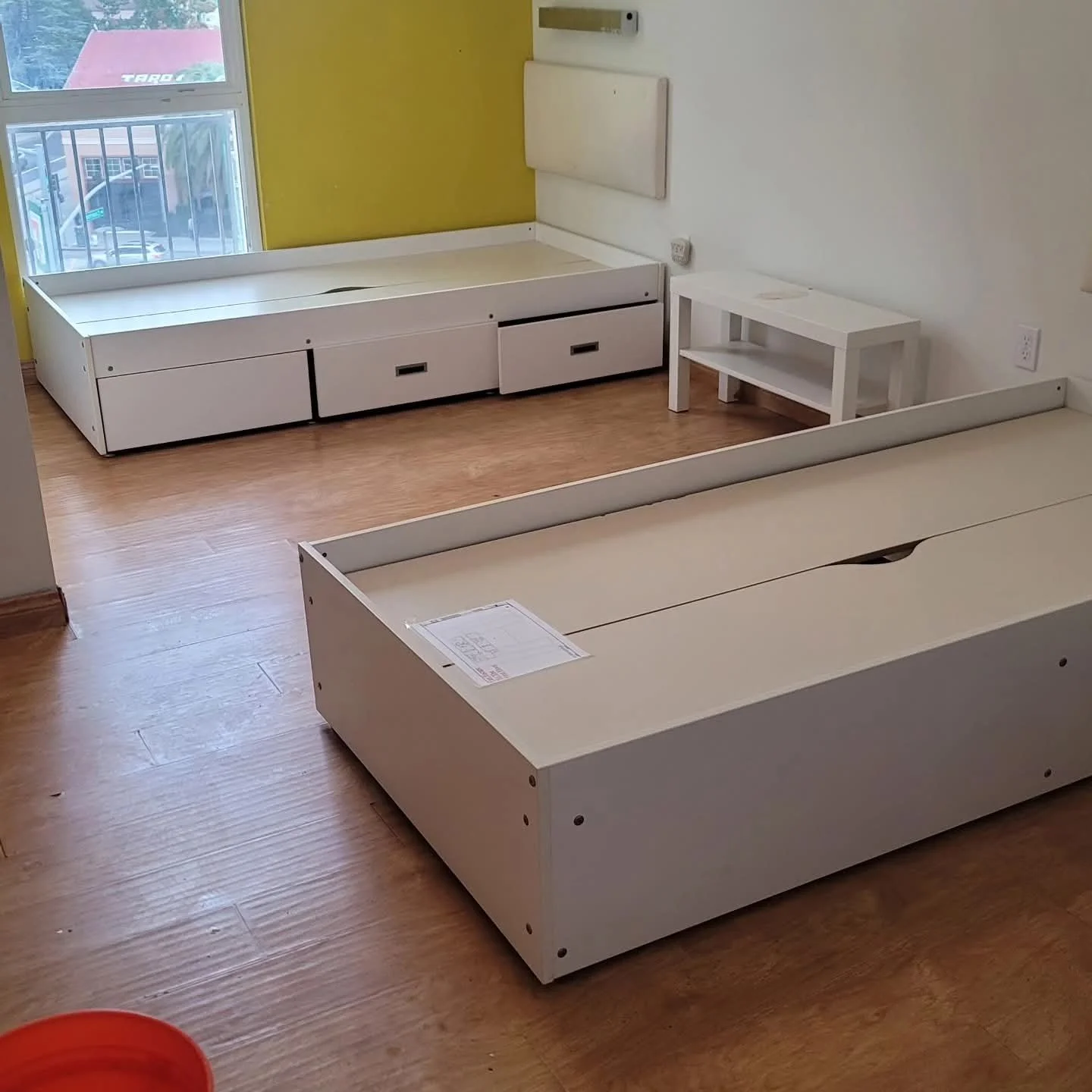 Empty white beds and furniture in a room with hardwood floors, a large window showing parked cars and buildings outside, and a yellow accent wall.
