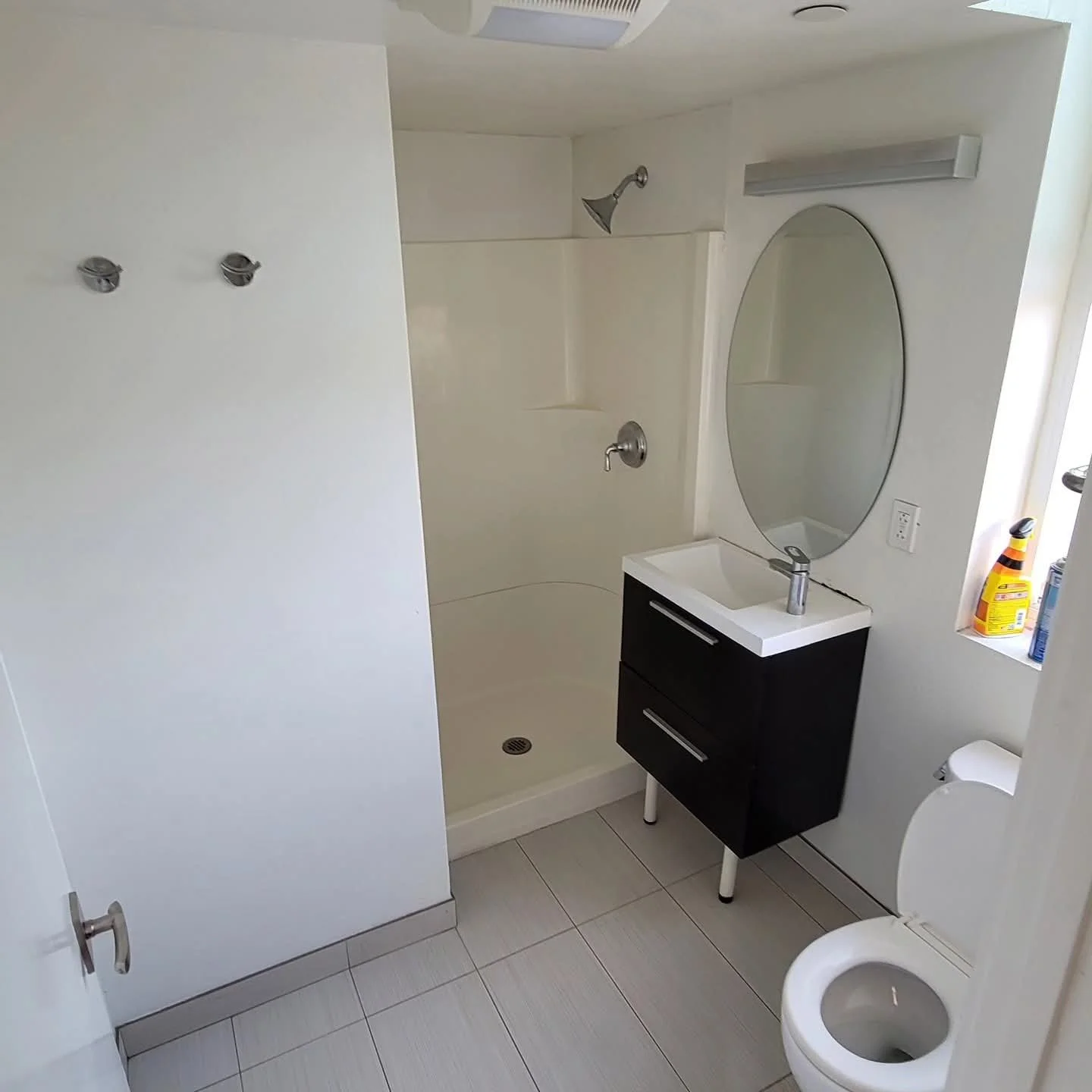 Small bathroom with a shower, sink with mirror, toilet, and cleaning supplies on window ledge.