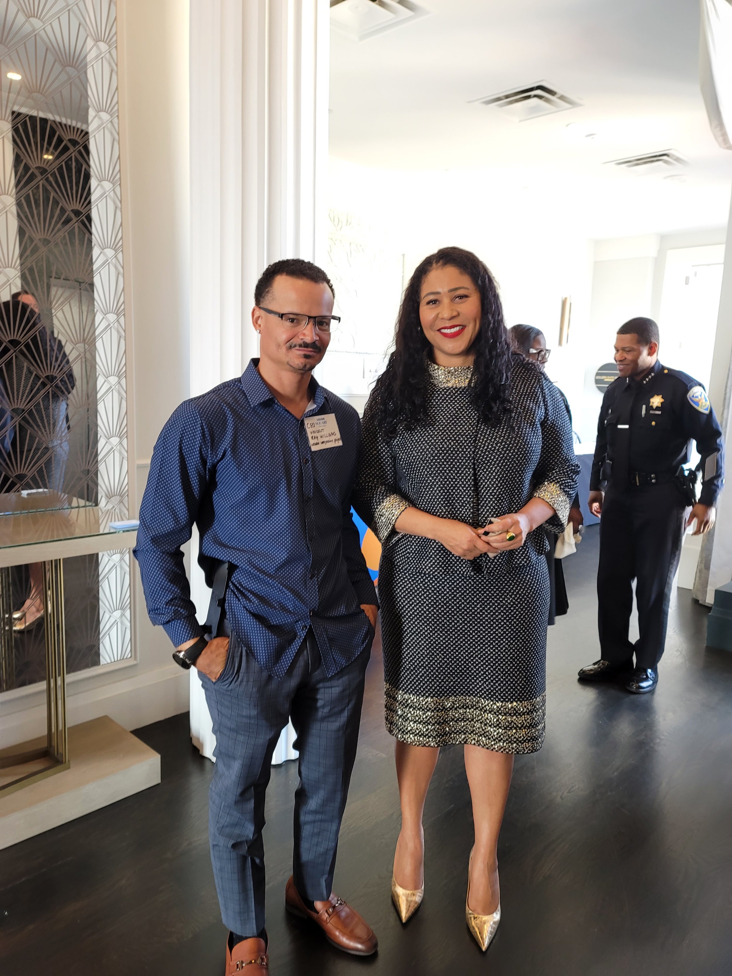 A man and a woman standing together indoors, smiling at the camera. The man is wearing a blue patterned shirt, gray pants, and brown shoes, with glasses and a name tag. The woman is wearing a black and white patterned dress, gold high heels, and a ring, with long curly black hair.