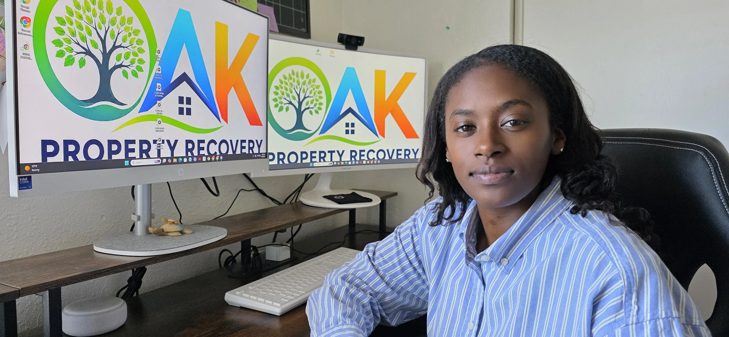 Carter Garden sitting at a desk with dual computer monitors displaying a logo with a tree, a house, and the words "OAK Property Recovery." She is wearing a blue striped shirt and has a neutral expression.