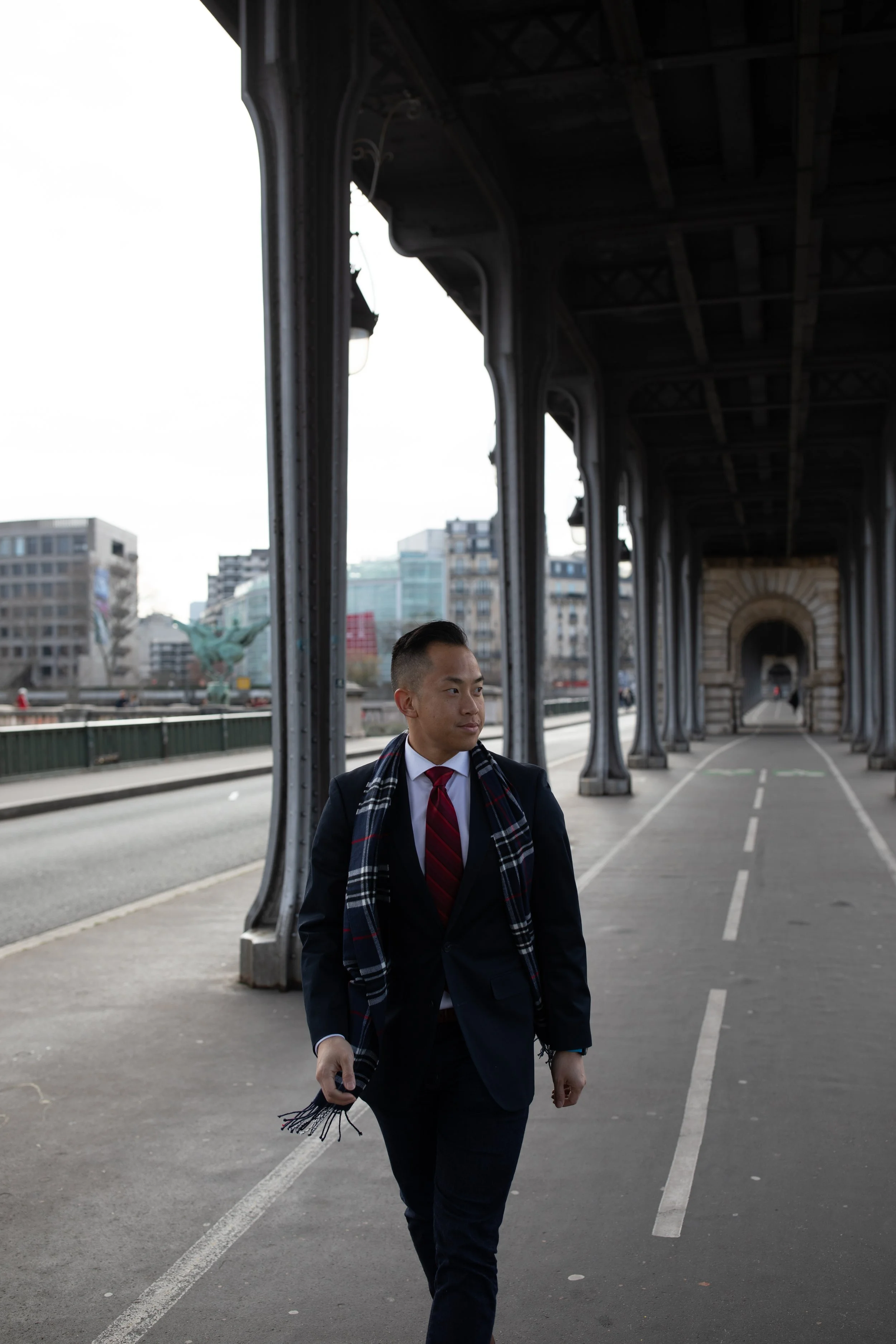 A young man in a suit and red tie walks along a city street under a bridge, carrying a scarf and a phone in his right hand.