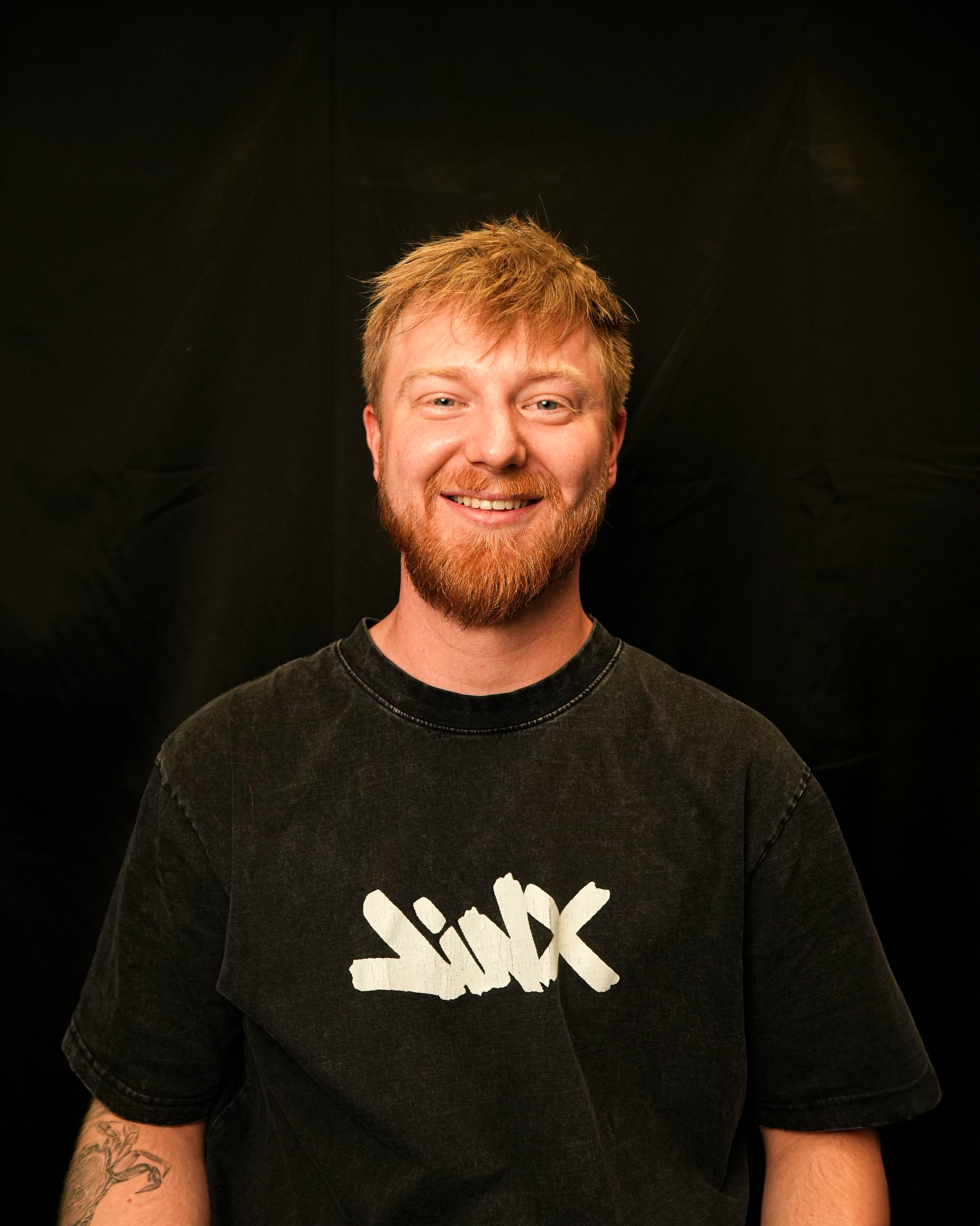 Portrait of a smiling man with red hair and beard wearing a black t-shirt with a white graphic, standing against a black background.