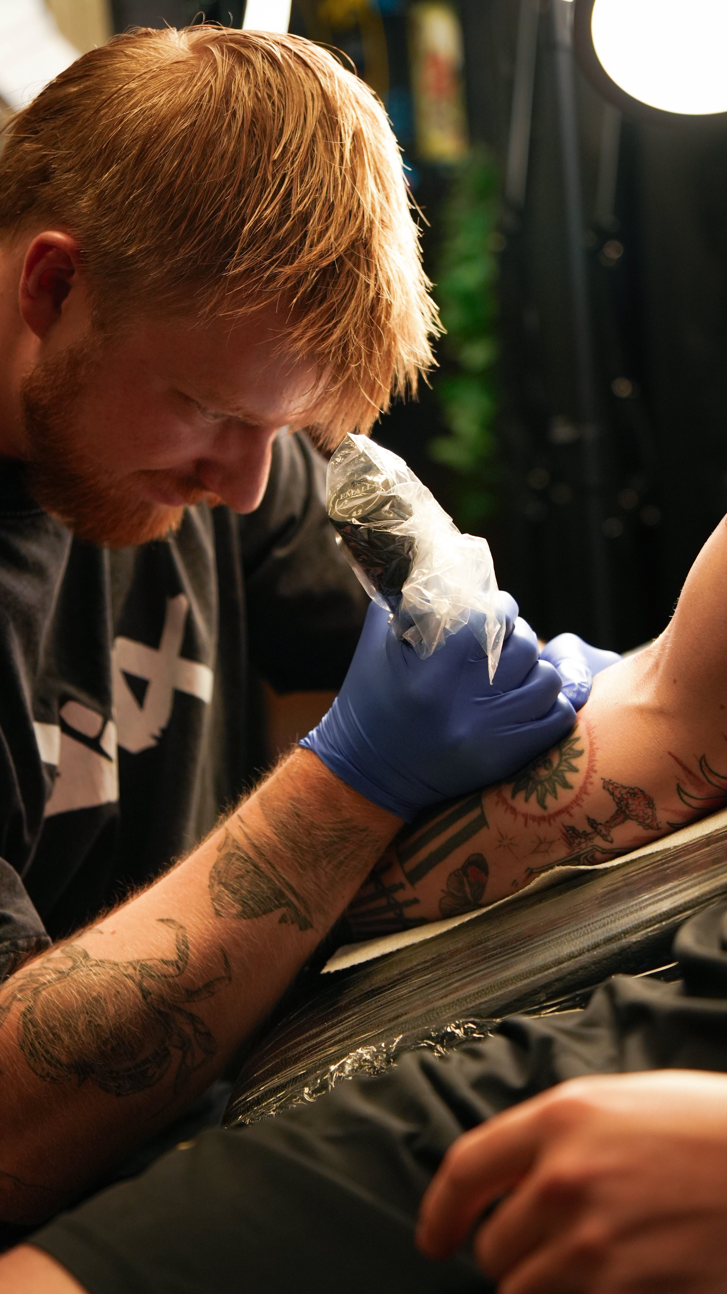 A tattooed person receiving a tattoo on their upper arm while a tattoo artist in gloves works on it with a tattoo machine.