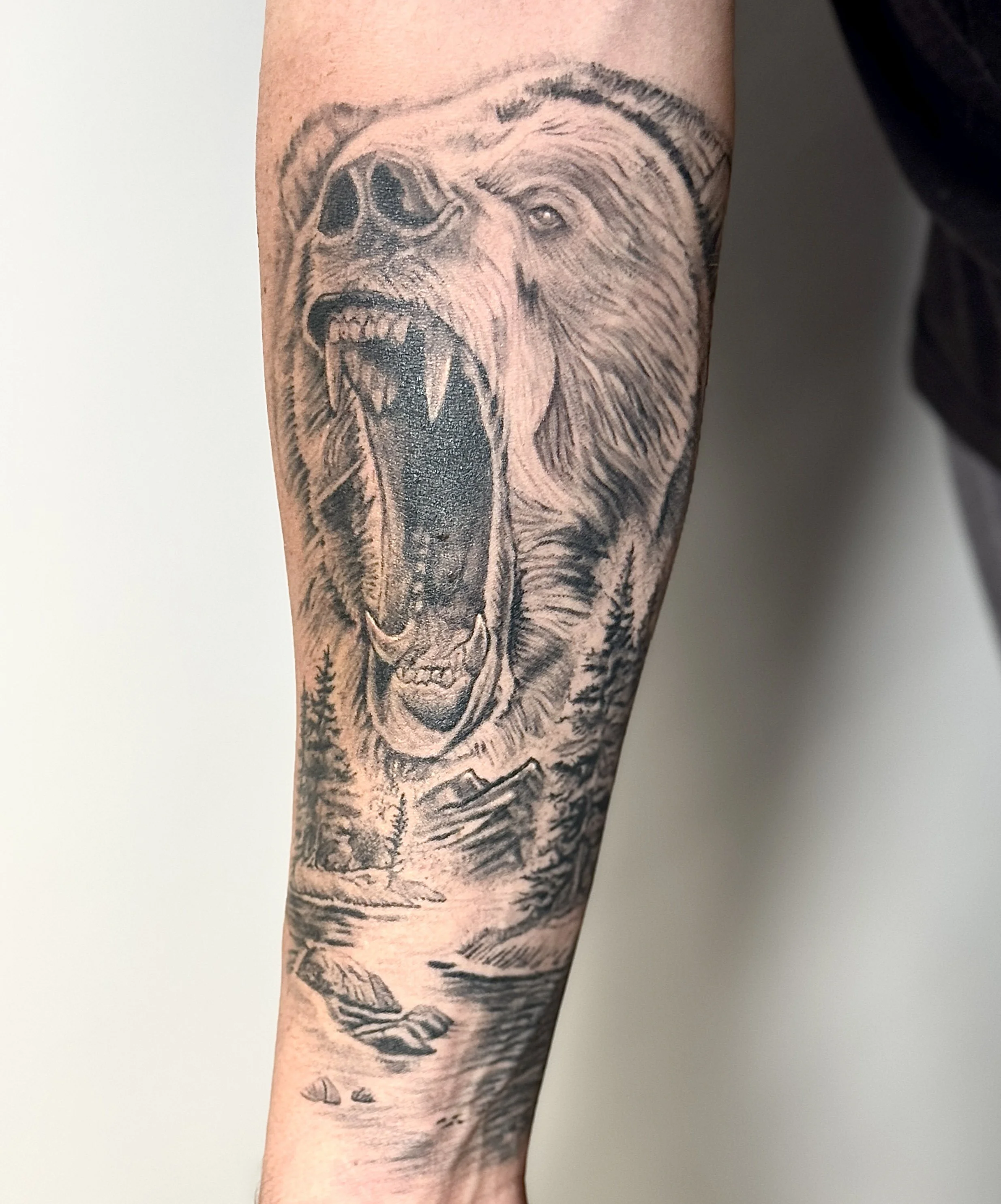 Black and gray tattoo of a roaring bear's face with detailed fur, nose, eyes, and open mouth showing teeth, on a person's arm.