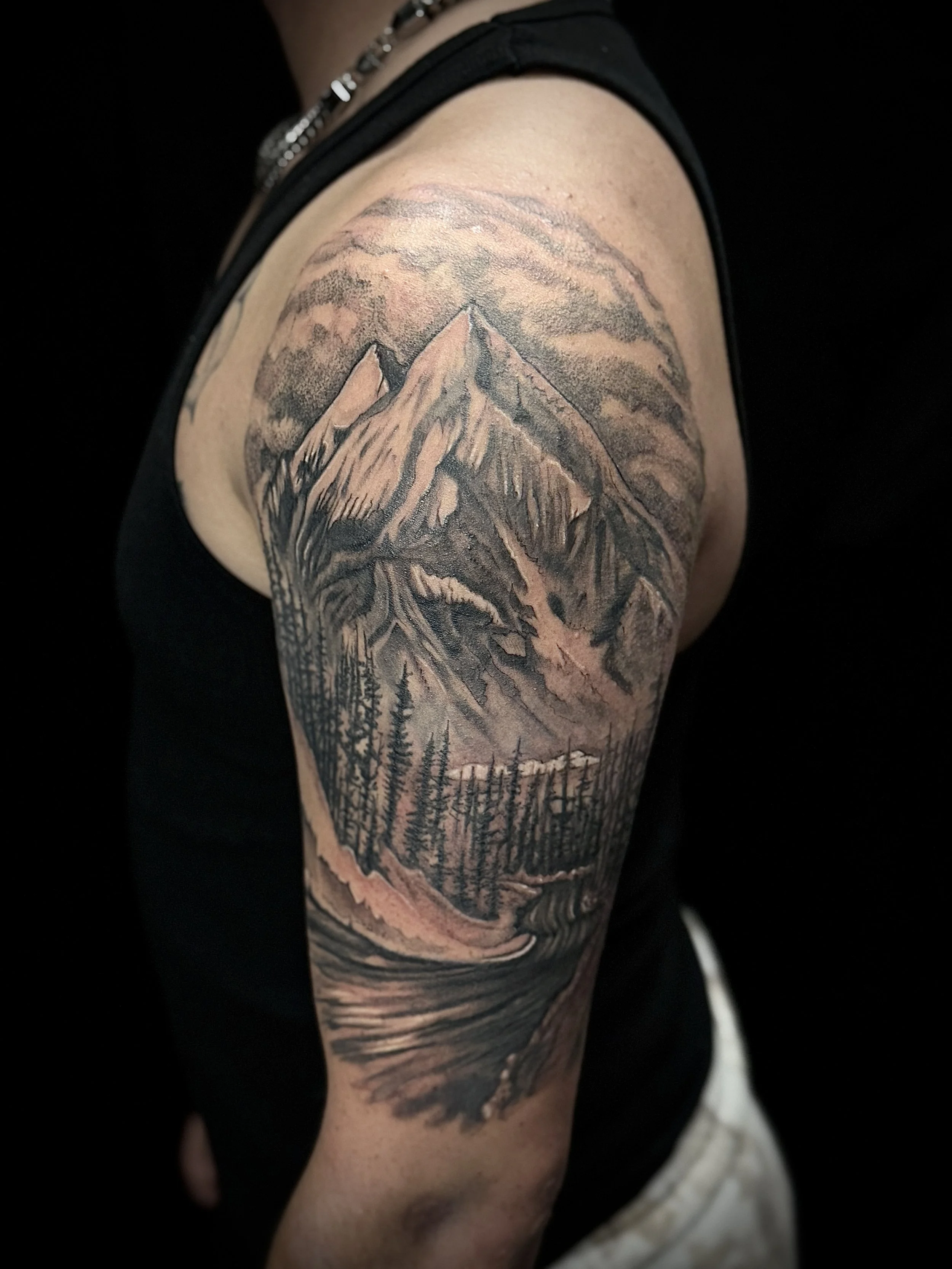 A tattoo of a mountain landscape on a person's upper arm, featuring a pine forest in the foreground and mountains in the background.