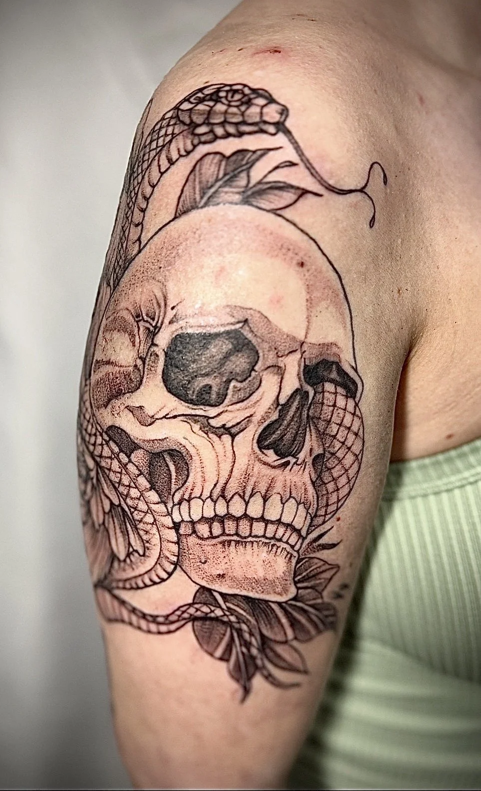 Tattoo of a skull with a snake wrapped around it, featuring leaves and decorative elements, on a person's upper arm.
