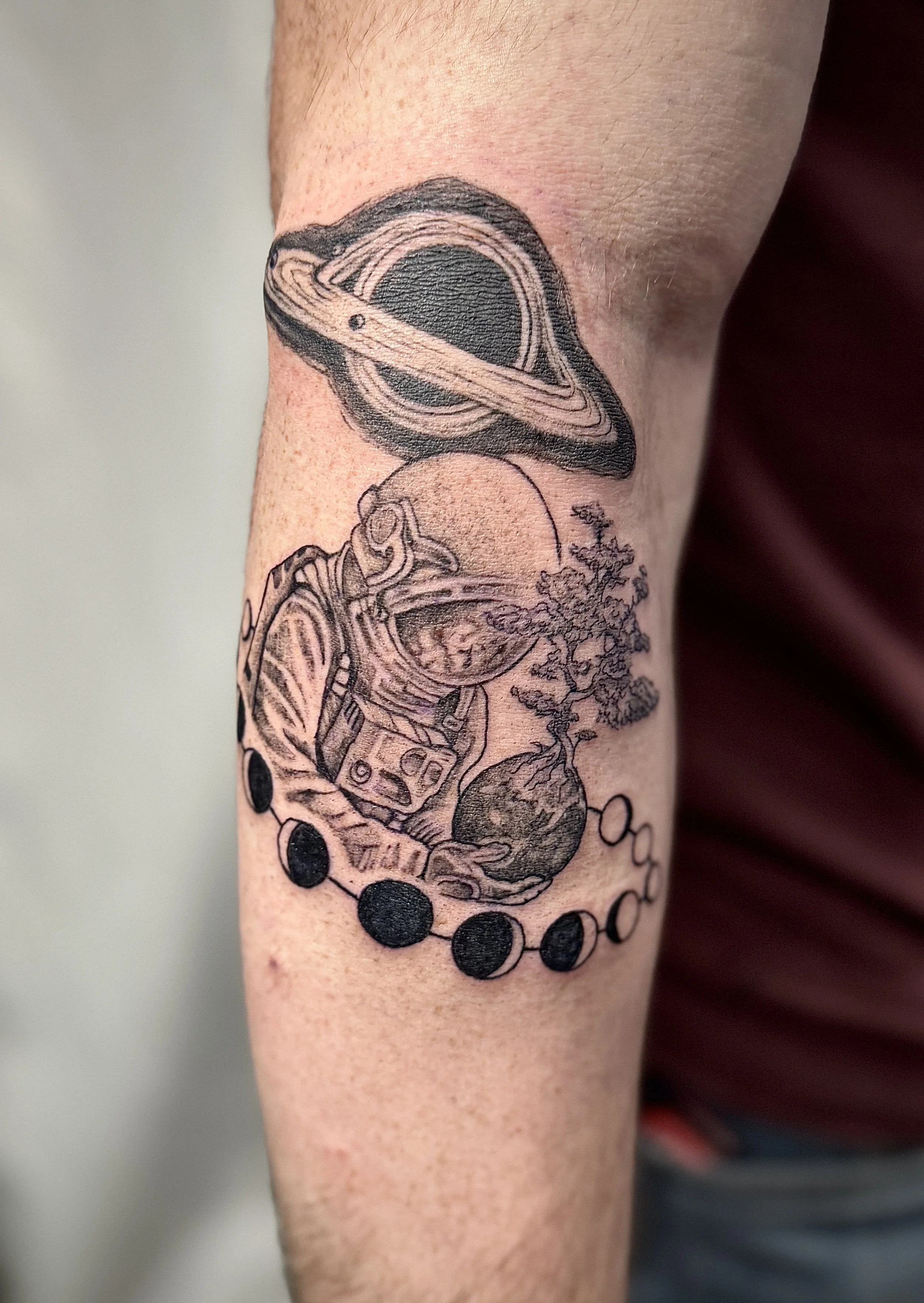 A tattoo on a person's forearm featuring a black and gray depiction of Saturn with detailed rings, and a space-themed design with planets, a tree, and a moon with craters.