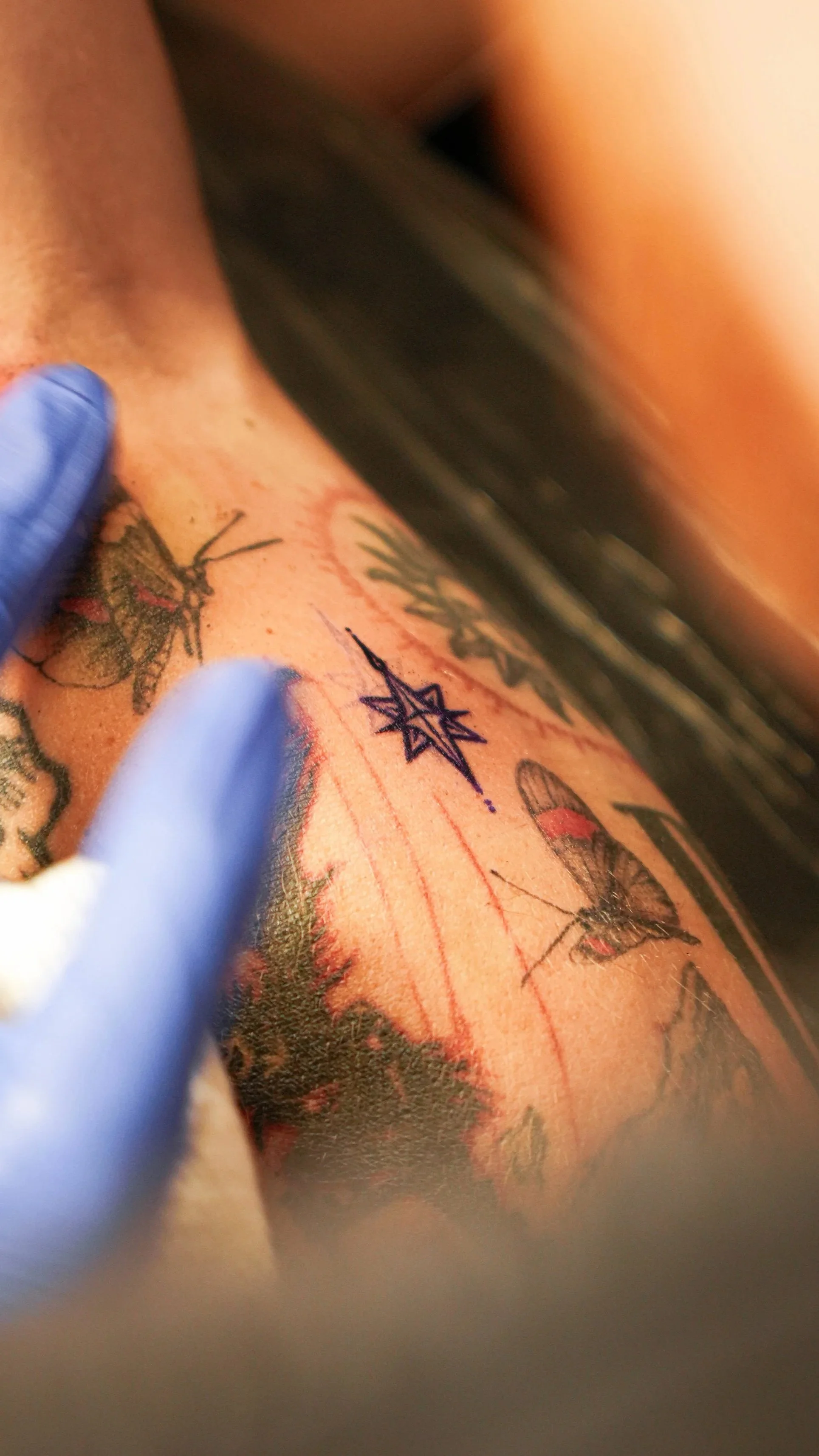 Close-up of a person's arm with several tattoos, including a bee, a star, and butterflies, while a person wearing blue gloves is performing a tattooing process.
