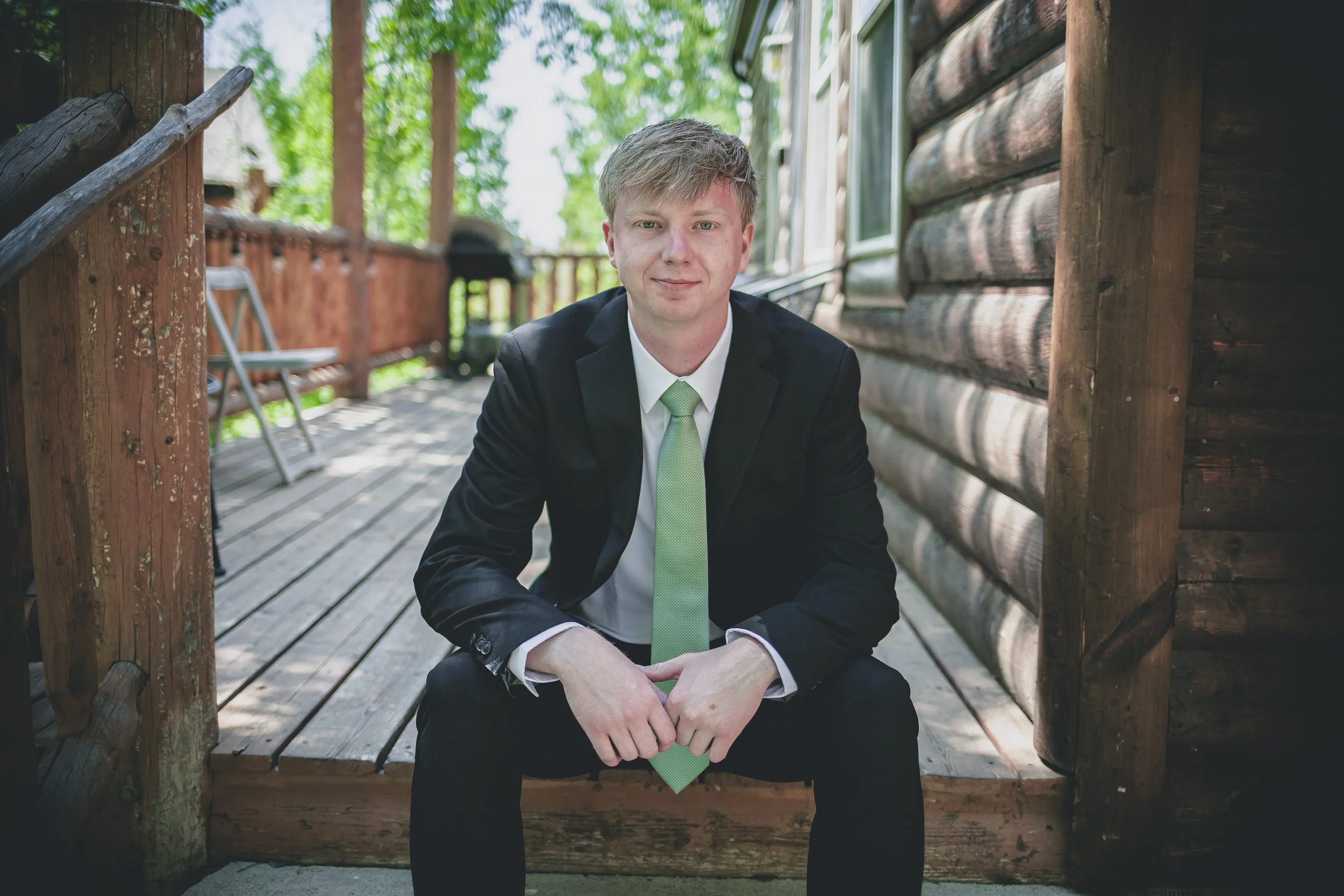 A young man in a business suit with a light green tie, sitting on a wooden porch with a log cabin behind him, smiling slightly at the camera.