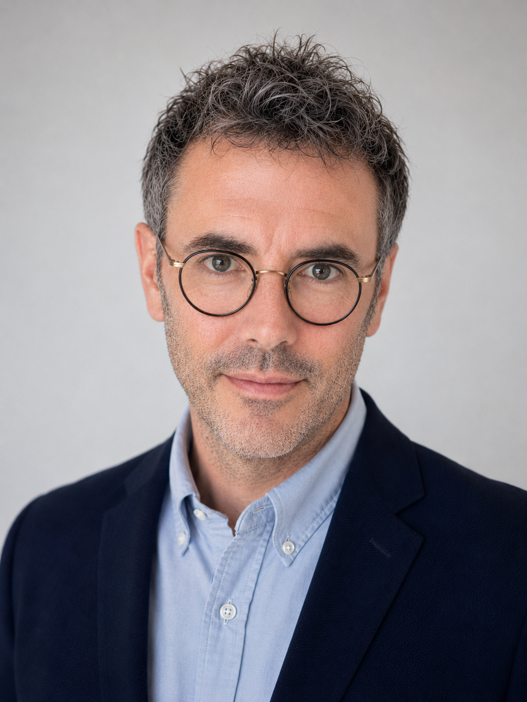 A professional headshot of a middle-aged man with gray hair and a beard, wearing round glasses, a dark blazer, and a light blue button-up shirt, against a plain background.