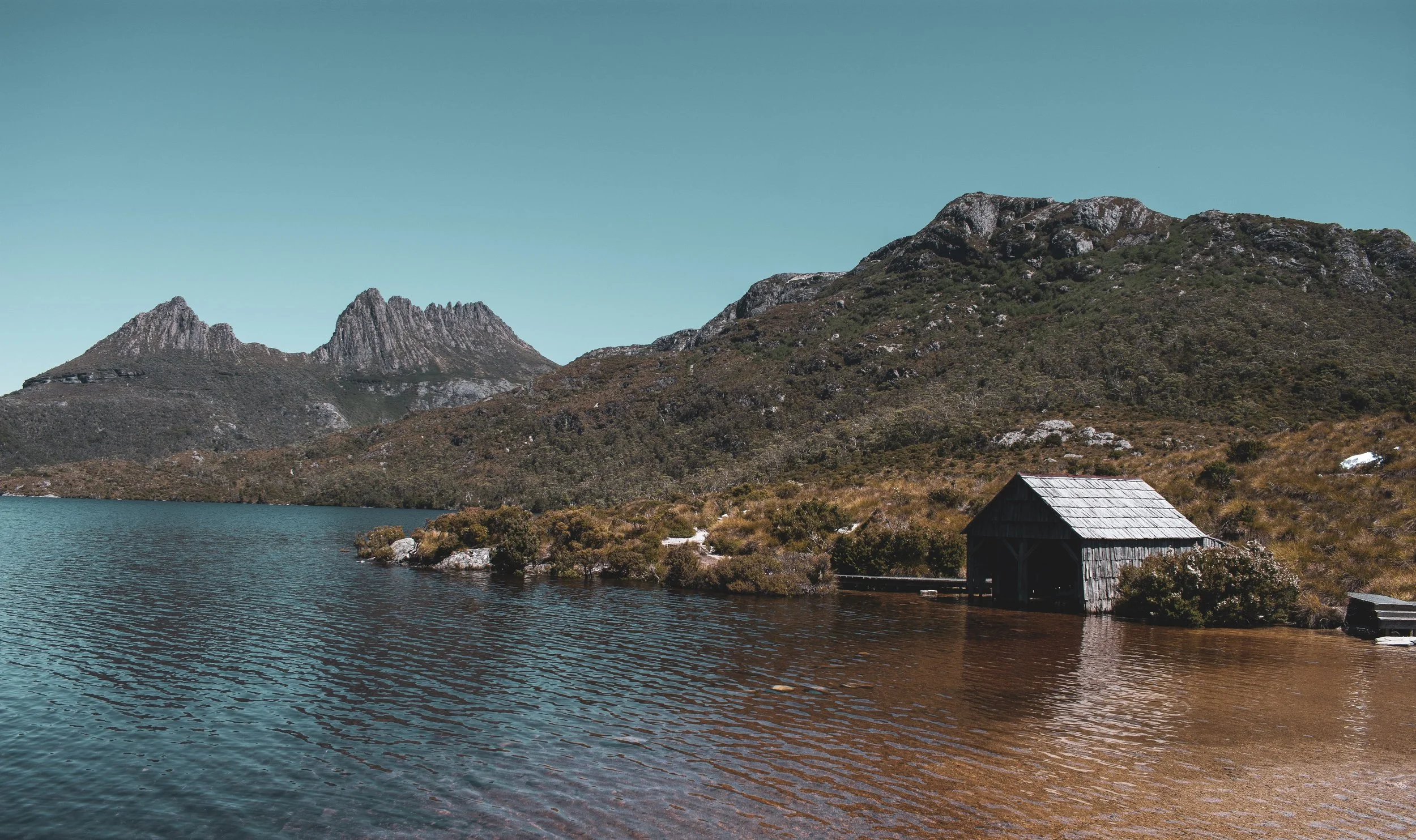 A peaceful mountain scene with a lake in the foreground, surrounded by rocky hills and a small wooden cabin near the water's edge, under a clear blue sky.