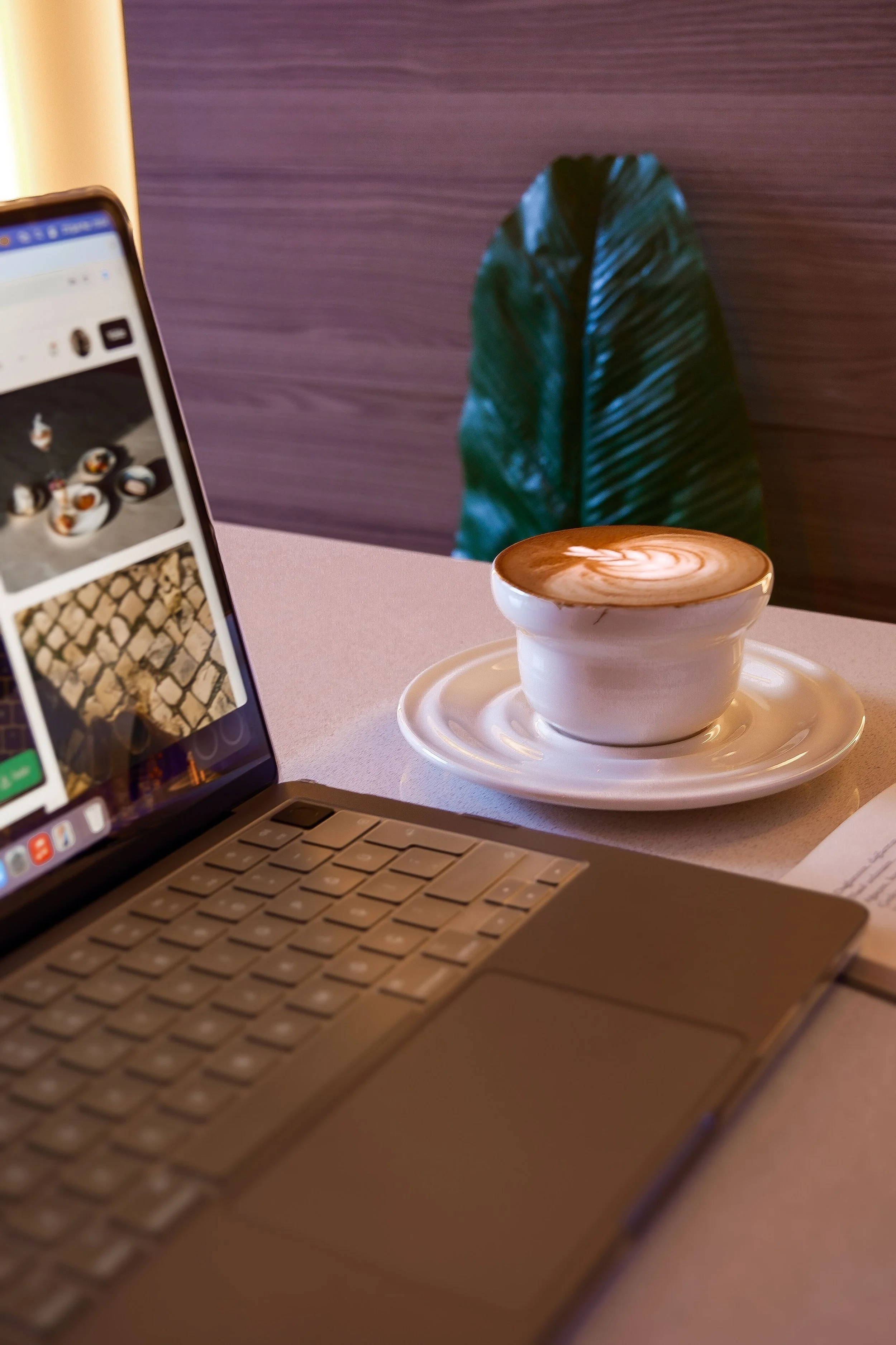Close-up of a silver laptop keyboard, a white coffee cup with latte art on a matching saucer, and a partially visible smartphone displaying a photo app with sushi images, on a pink surface, with a large green leaf and purple wall in the background.