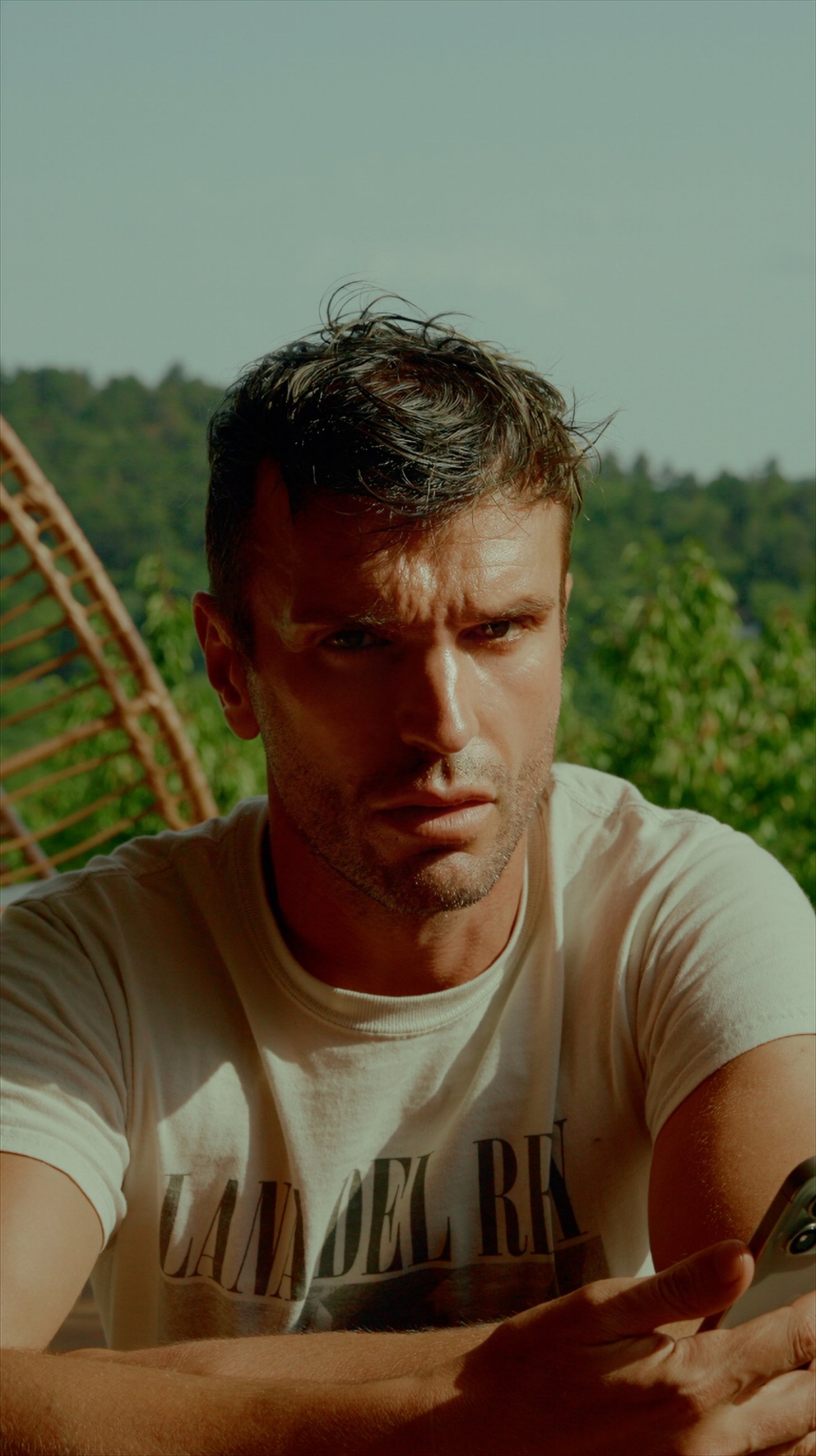 A man with short dark hair and a serious expression looking at a camera, wearing a white t-shirt, with a natural outdoor background and greenery.