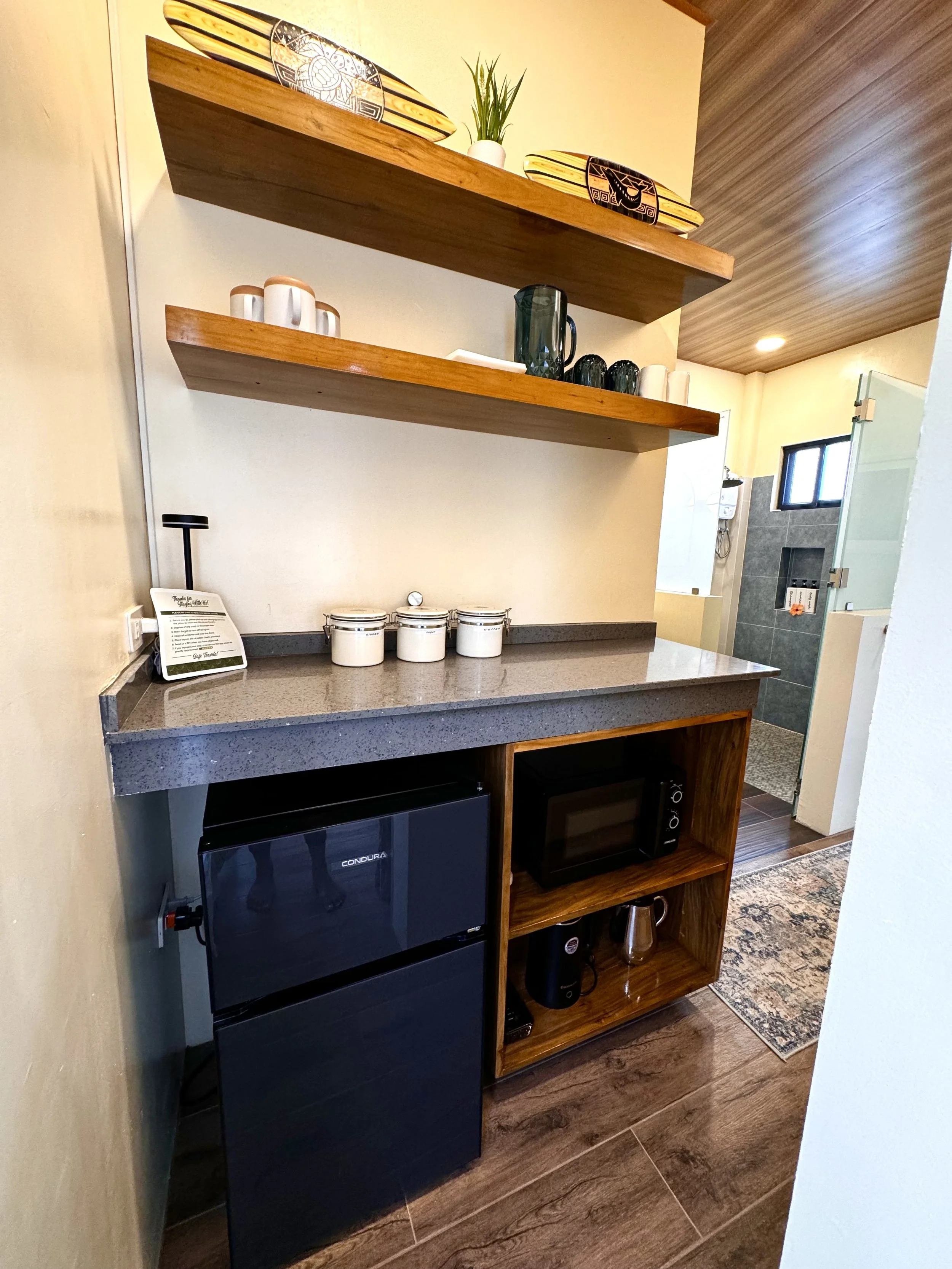A kitchenette area with open wooden shelving holding decorative bowls, a small plant, and cups. Below are jars, a microwave, and shelves with a coffee machine and cups. The floor is wooden, and there's a partial view of a bathroom with a window, tile