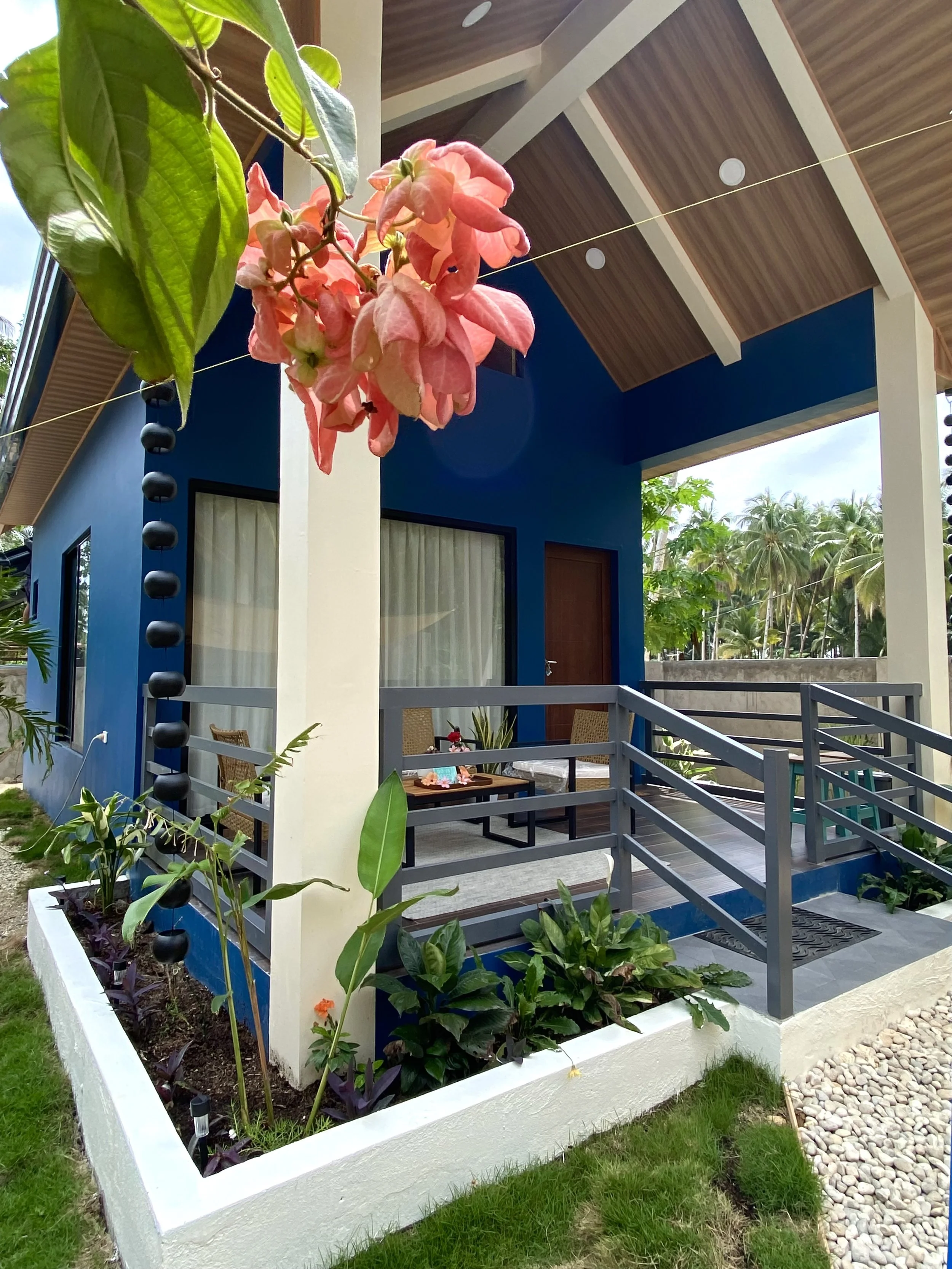 A blue house with a porch, featuring a garden with green plants and purple-leafed plants, a small table with chairs, and a gravel pathway bordered by a white wall.