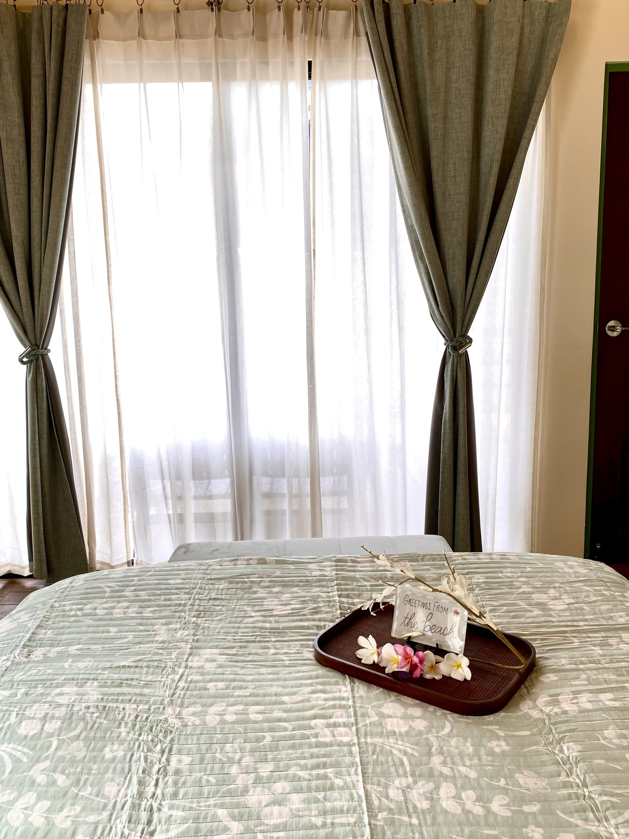 A curtained window with sheer white curtains and dark green curtains tied back. Part of a bed with a light-colored patterned bedspread is visible in the foreground. On the bed, there's a wooden tray with white and pink flowers and a small sign that r
