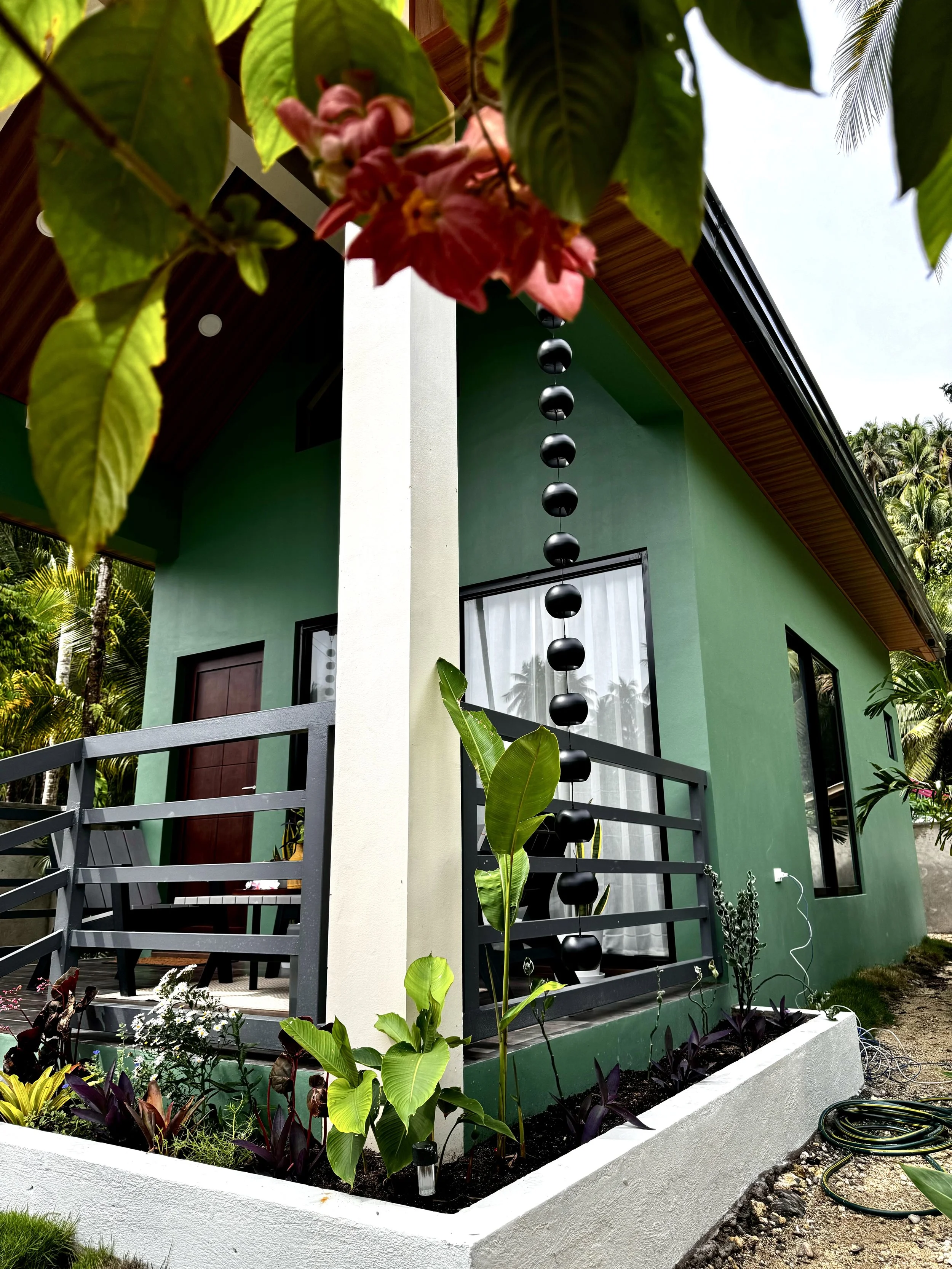 A small garden with various plants and flowers, enclosed by a white painted concrete border, adjacent to a modern green house with large windows, a decorative rain chain hanging from the roof, and a deck with a black railing.