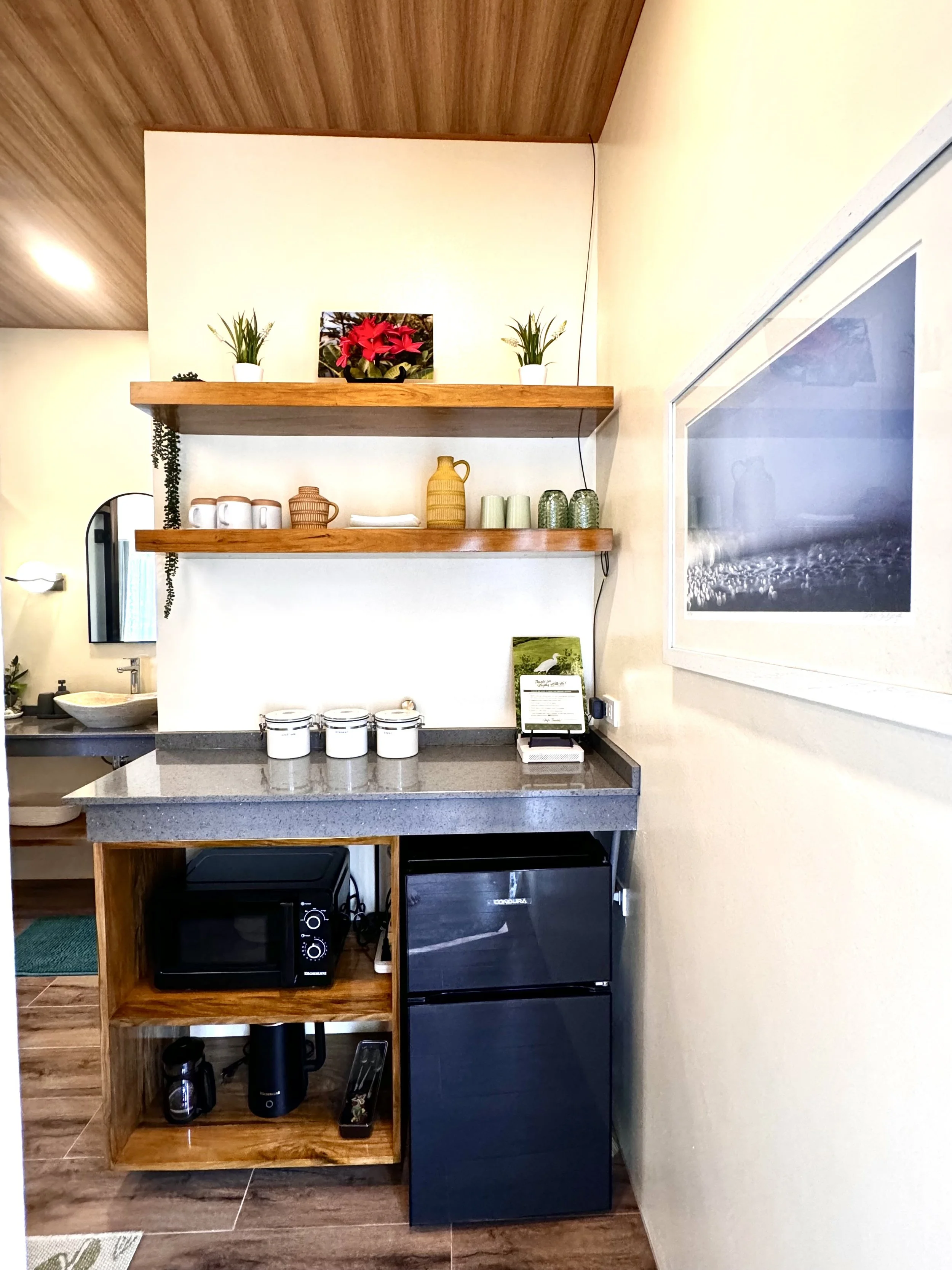 Wooden open-shelving unit with kitchen appliances and decor, including a microwave oven, a small black refrigerator, white mugs, a woven jug, a yellow pitcher, green cups, and potted plants, set against a white wall with framed artwork.
