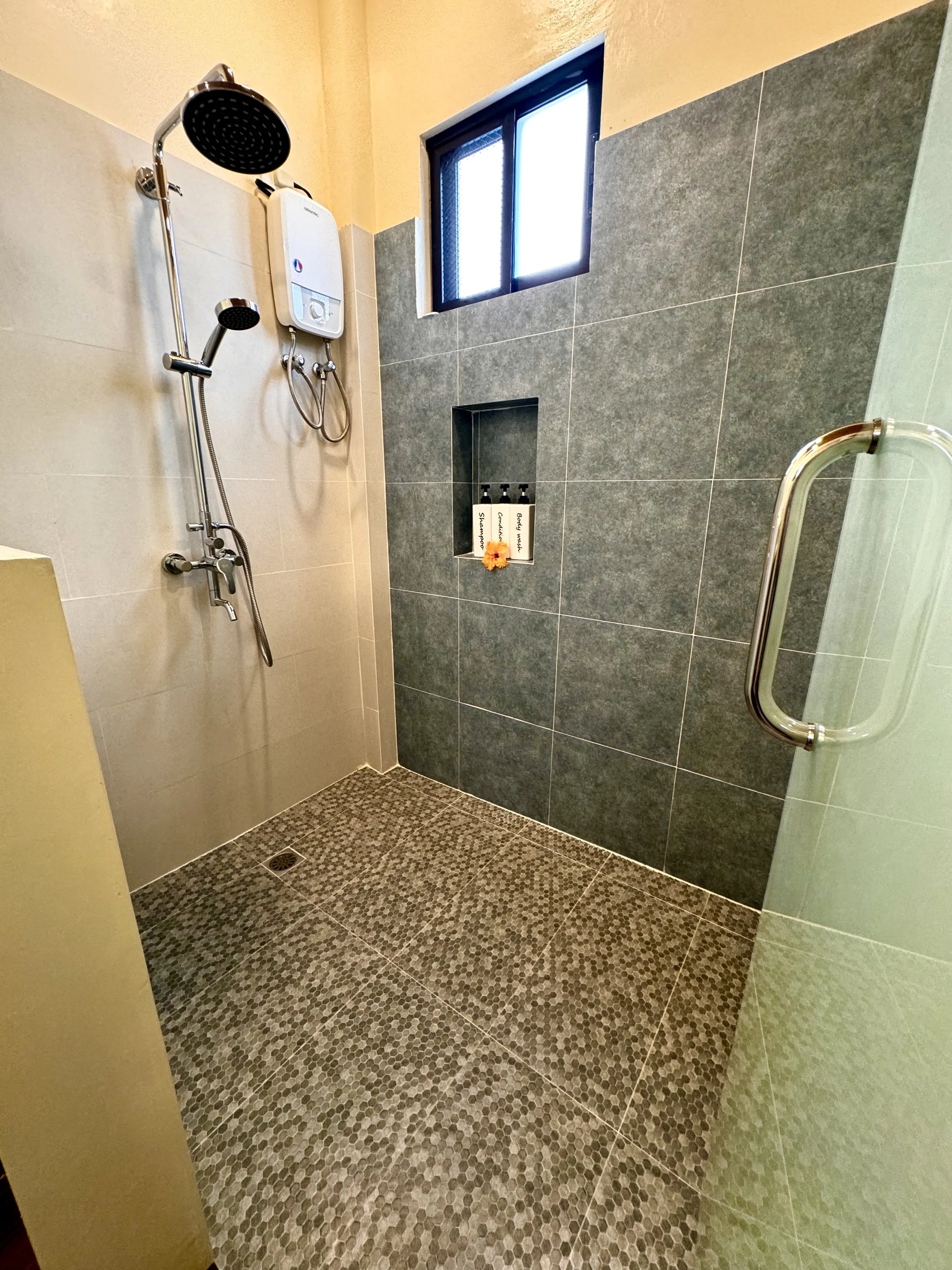 A shower room with grey and beige tiles, a window, a handrail, and bottles of shampoo, conditioner, and body wash.
