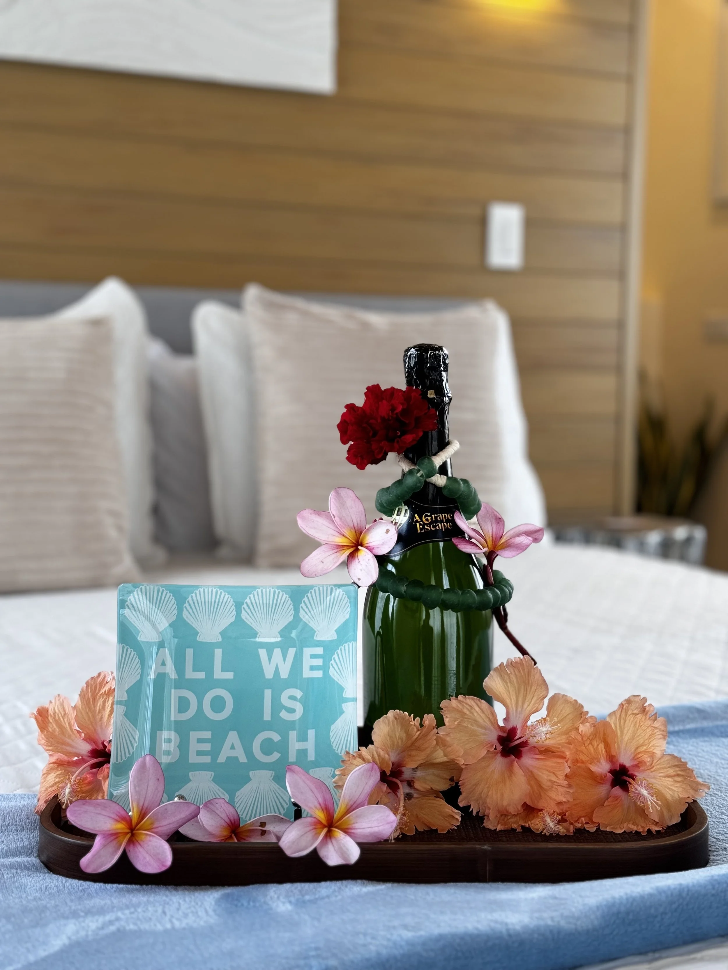 A tray with a bottle decorated with a red flower, surrounded by pink and orange flowers, a blue card with white text, and a background of a bed with multiple pillows and a wooden wall.