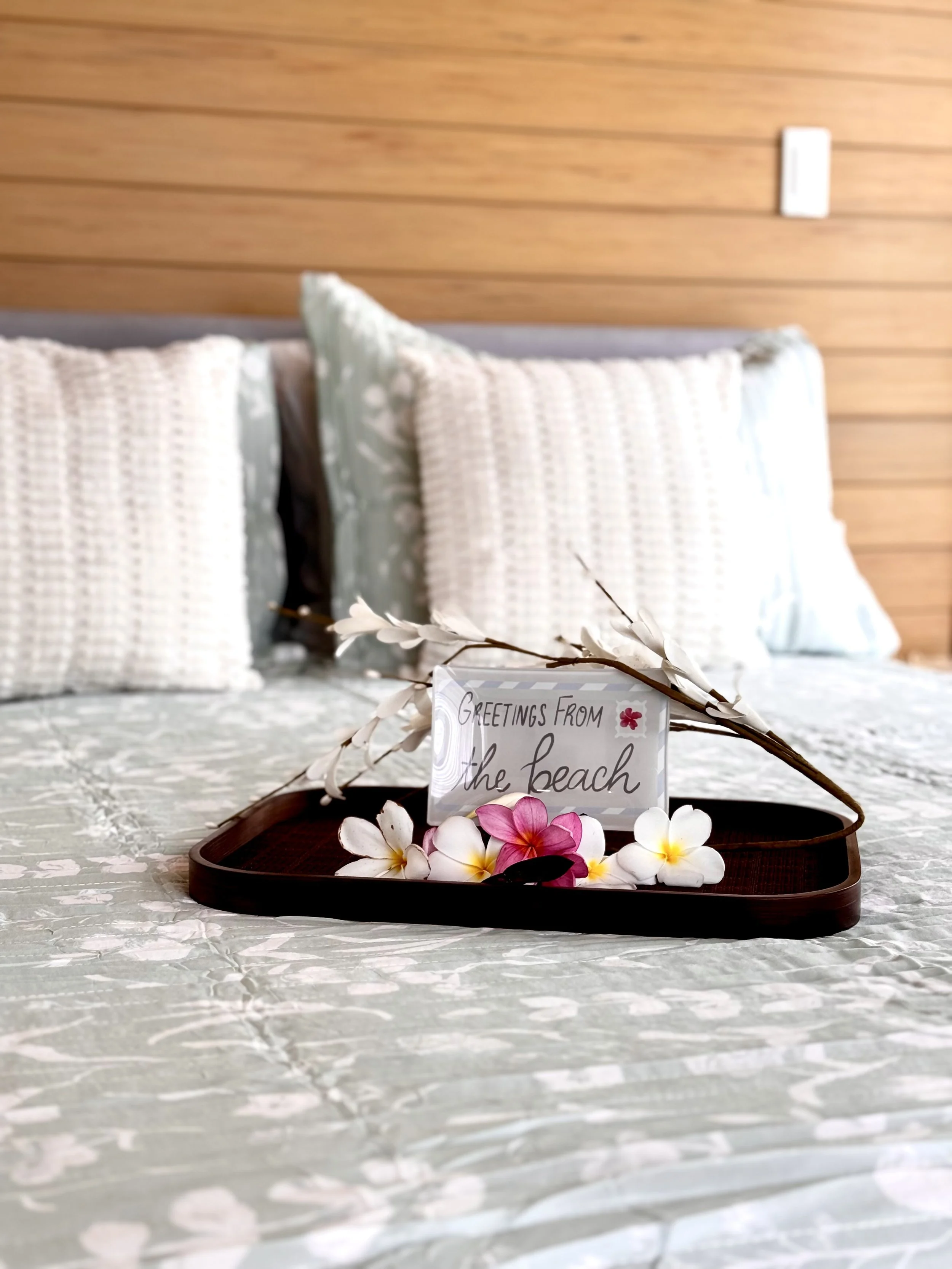 A decorative tray with pink and white flowers, a branch with white flowers, and a card that reads 'Greetings from the beach' set on a table with a textured tablecloth, with pillows and a wooden wall in the background.