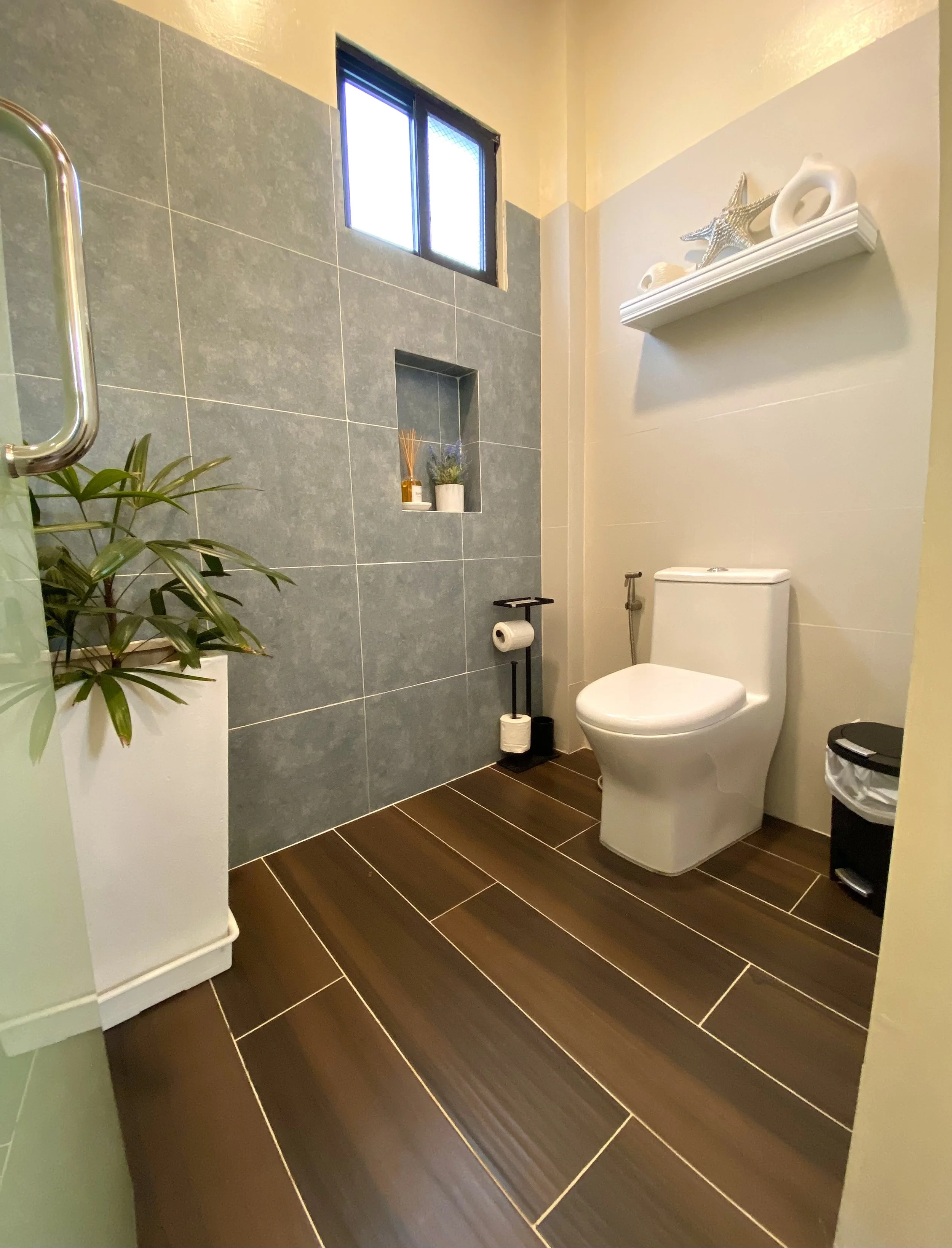 A modern bathroom with dark brown wooden floor tiles, a white toilet, a wall shelf with decorative seashell and starfish sculptures, a small recessed shelf with plants, a toilet paper holder, and a window with blue tint in the upper corner.