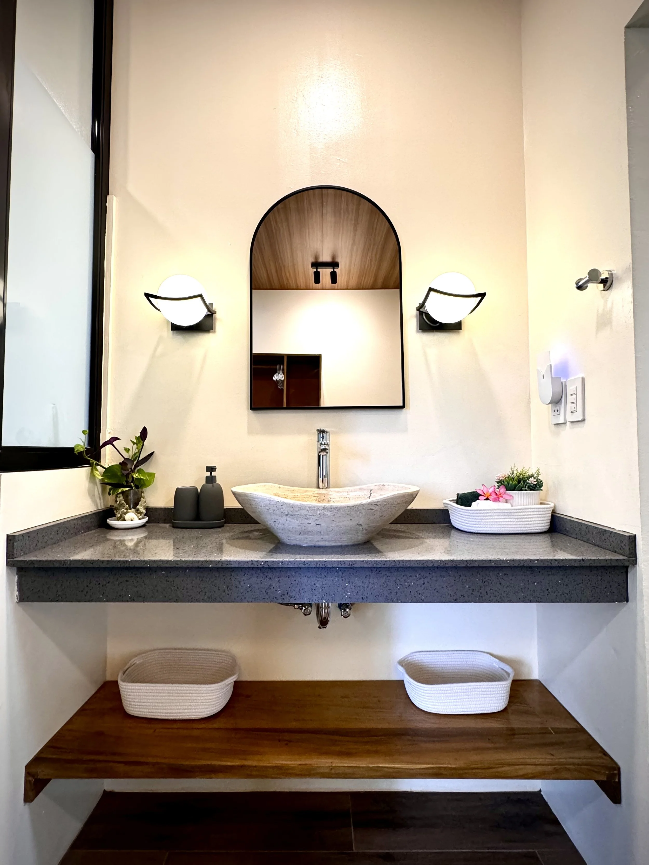 A bathroom vanity with two white baskets underneath, a stone sink on a gray countertop, a mirror with a wooden arch above, two wall-mounted lights on either side, a small plant, soap dispenser, and decorative items on the countertop, with a window to