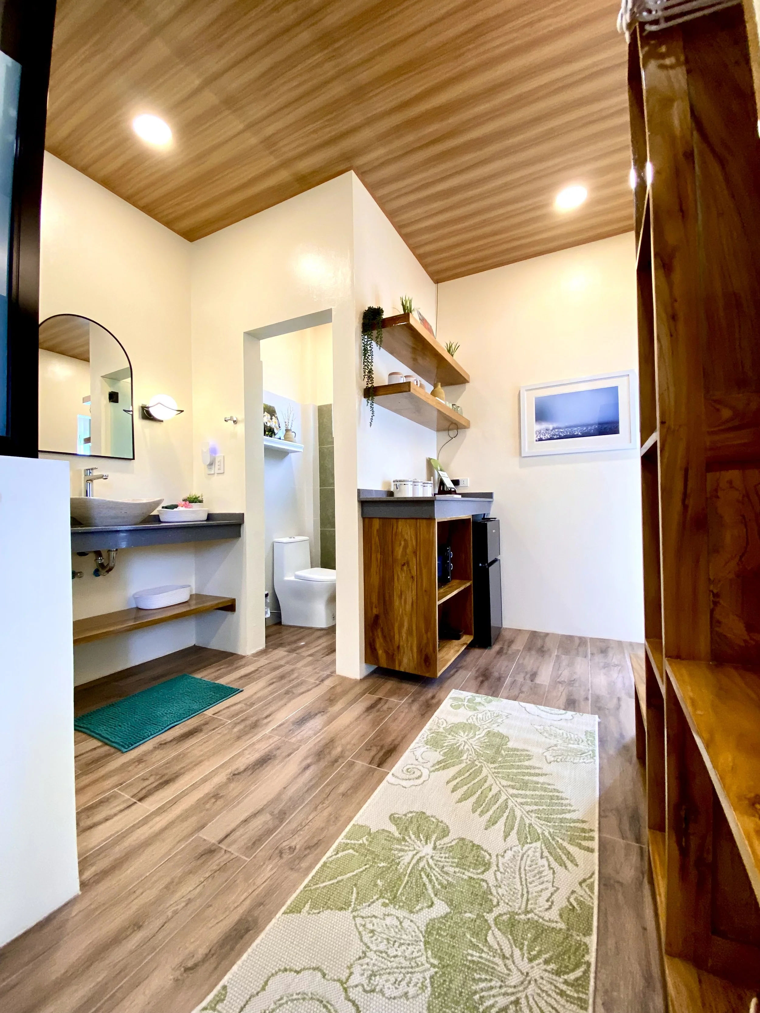 View of a cozy living space with wood flooring, a wooden ceiling, a small sink area with a mirror, a bathroom with a toilet, a wooden bookshelf, and decorative plants and paintings.