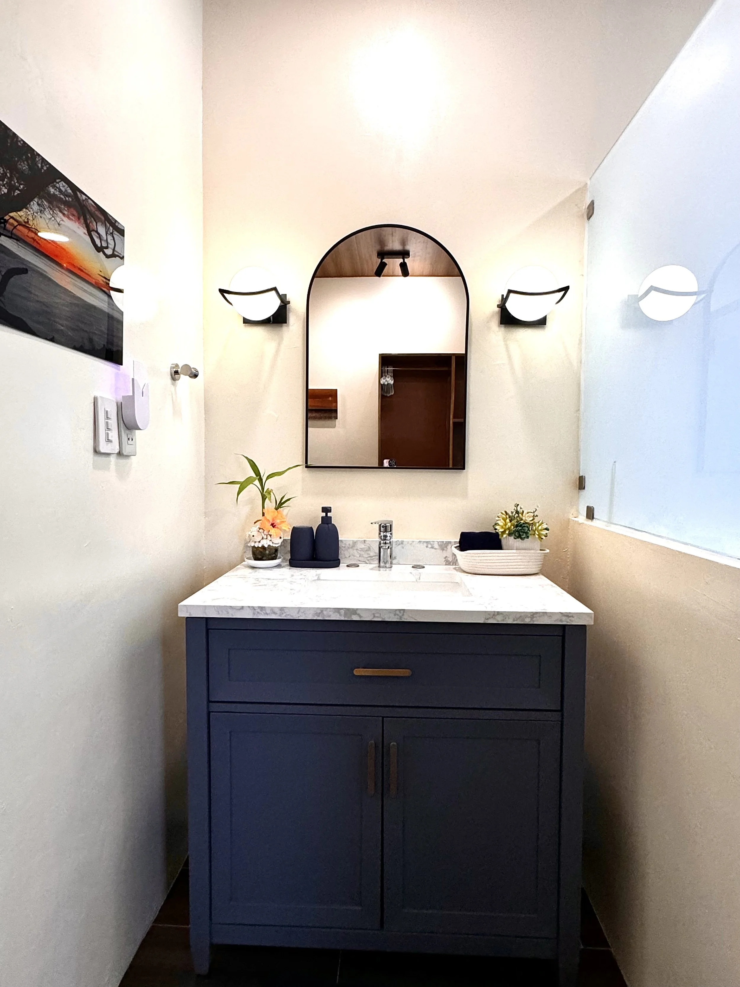 Bathroom vanity with a dark blue cabinet, a white marble countertop, a mirror, and decorative plants and bottles on the counter.