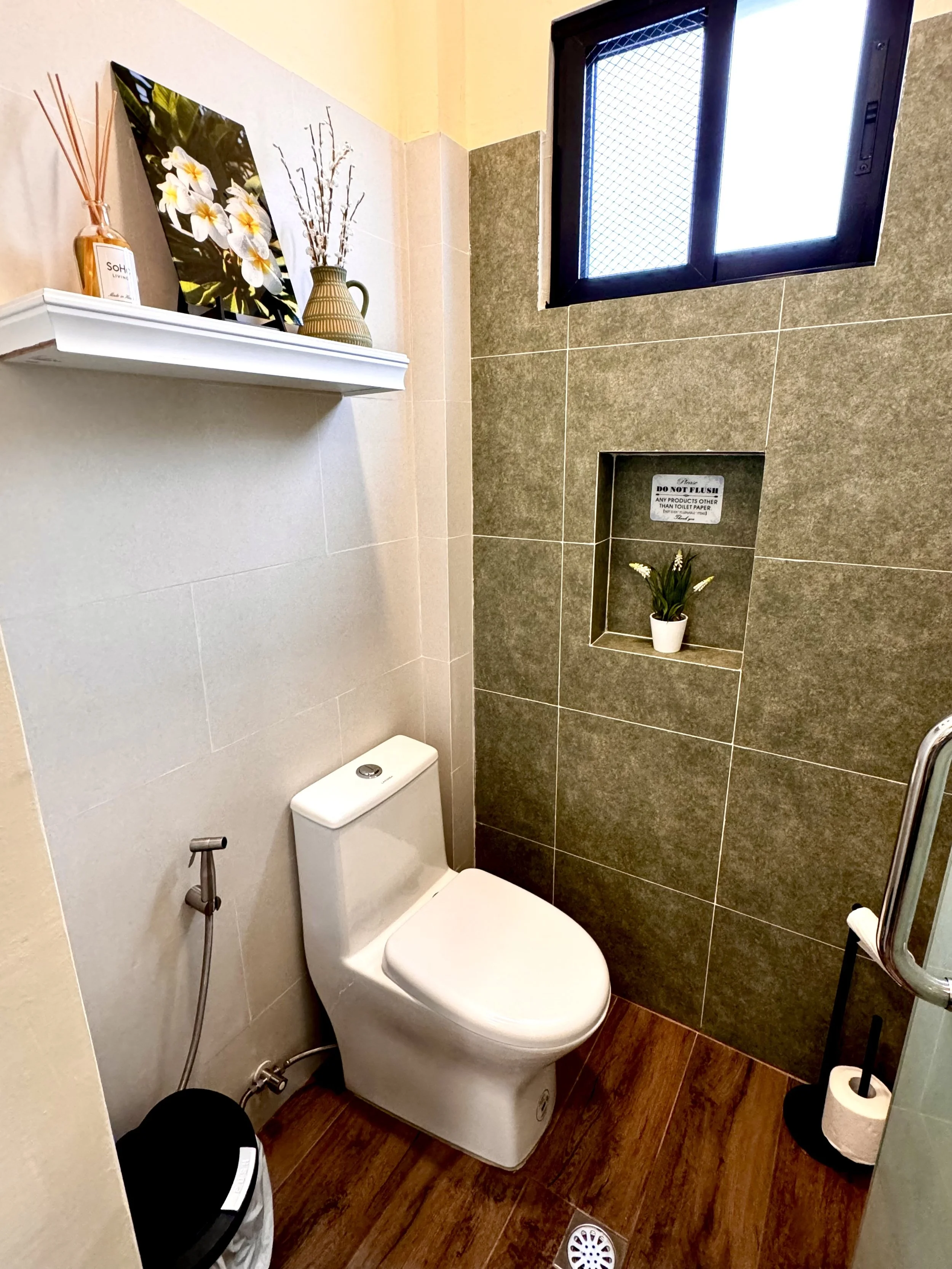 A small bathroom with a white toilet, a handheld bidet, and dark brown wooden flooring. The wall behind the toilet has beige tiles while the other wall has darker brown tiles with a built-in shelf holding a small potted plant. There is a small window