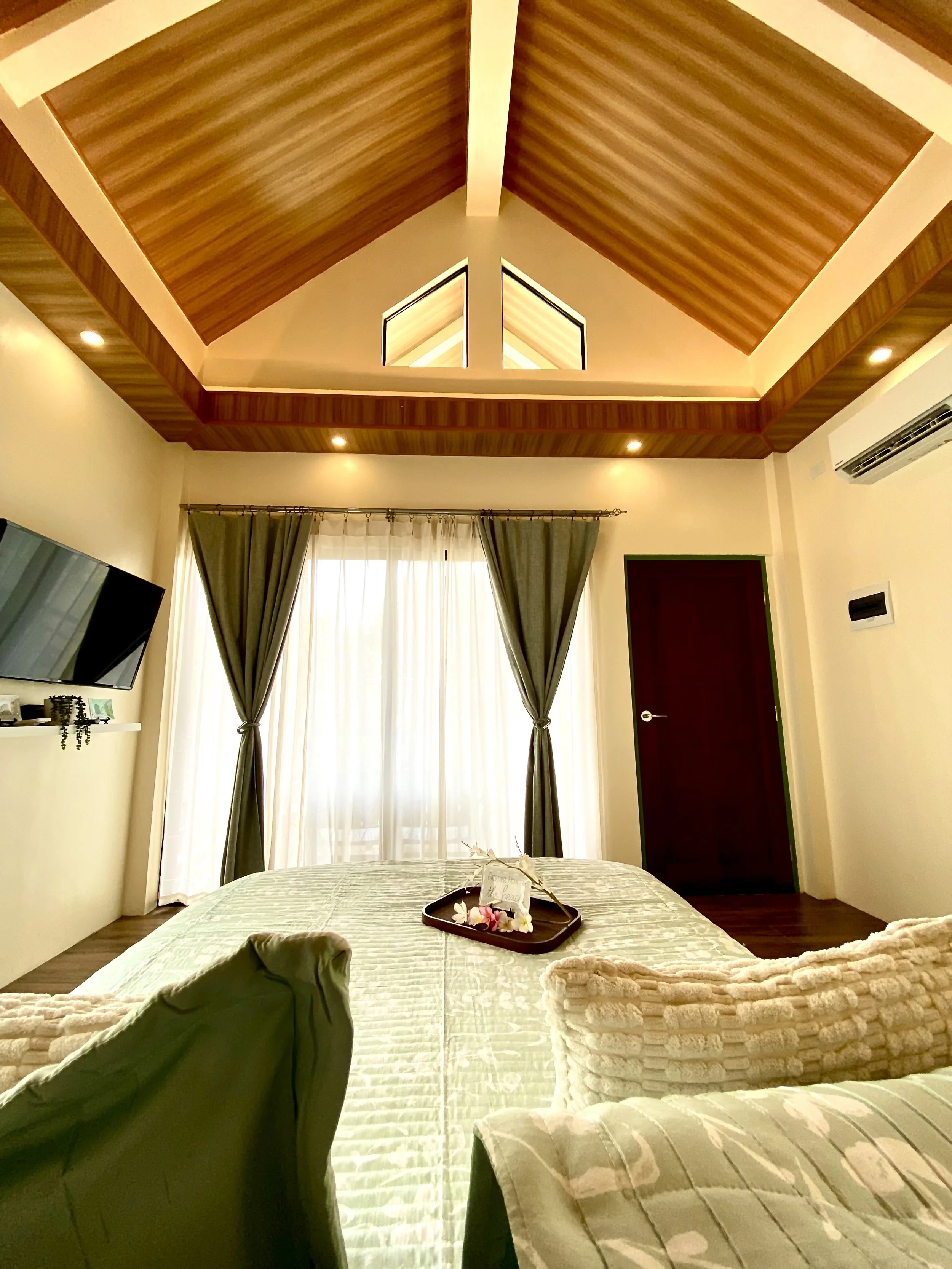 A bedroom with a vaulted wooden ceiling, white walls, and large window with sheer white curtains and green drapes. There is a bed with a tray holding a card and flowers, and folded towels on the bed. A TV is mounted on the wall, and an air conditioni