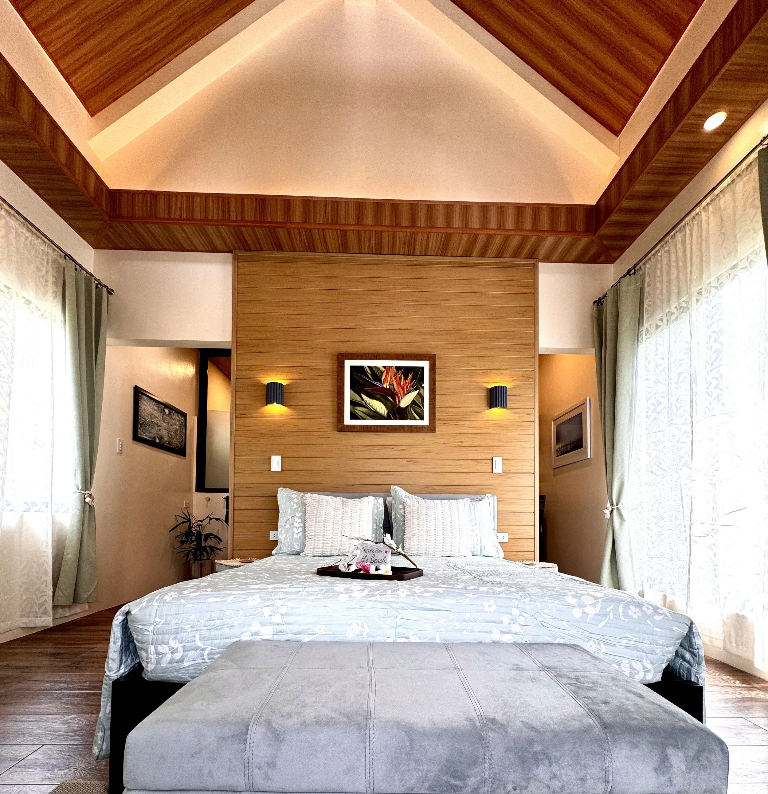 A bedroom with a bed, decorative pillows, a tray with floral decorations, and wooden paneling on the wall behind the bed. Curtains are seen on both sides of the window, and the ceiling has a cove lighting feature with wood accents.