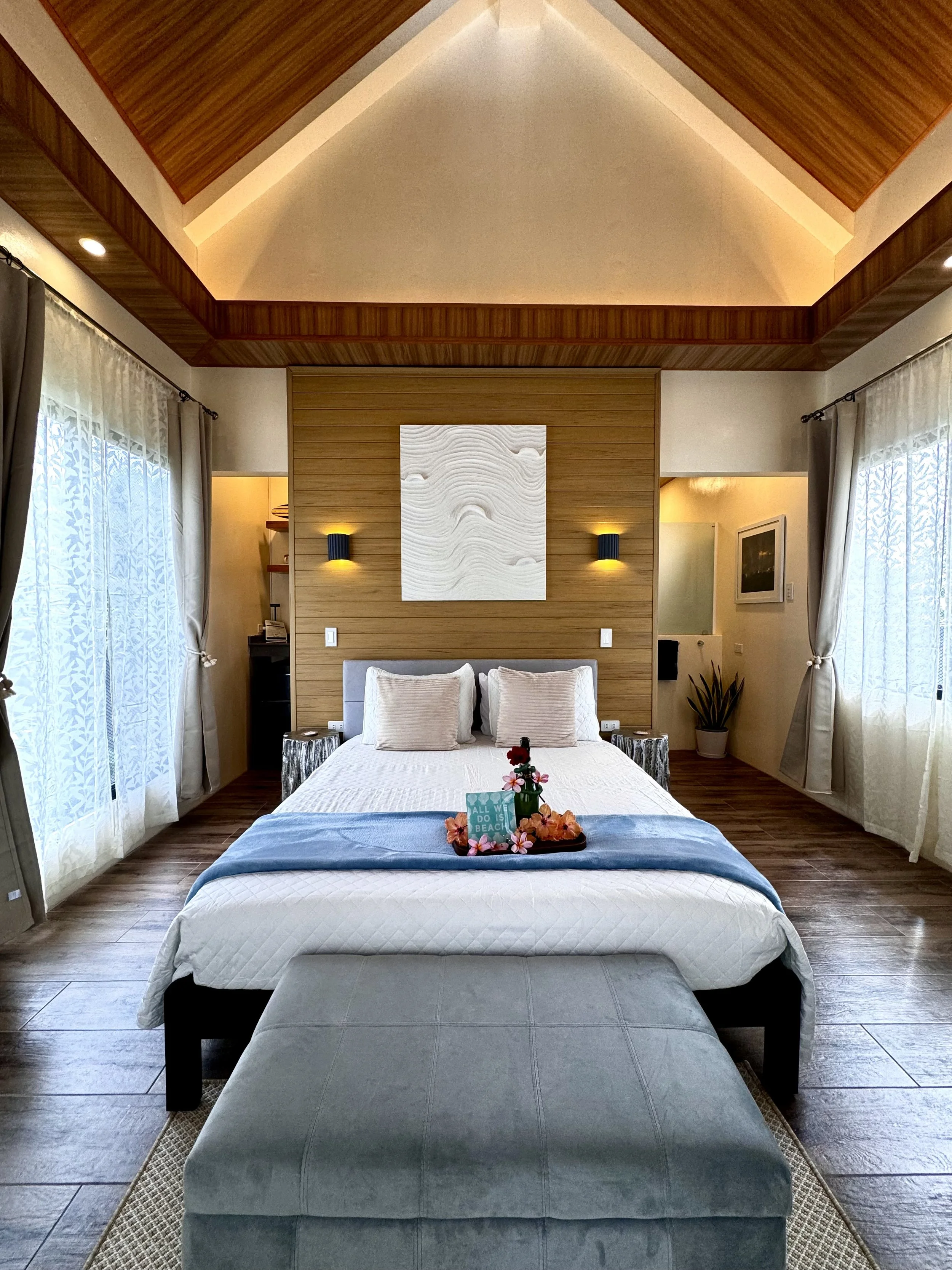 A neatly made bed with a white quilt, beige pillows, and a blue throw blanket, with a wooden headboard featuring two yellow wall sconces, in a bedroom with large windows with curtains, and a tray with flowers and a small sign on the bed.
