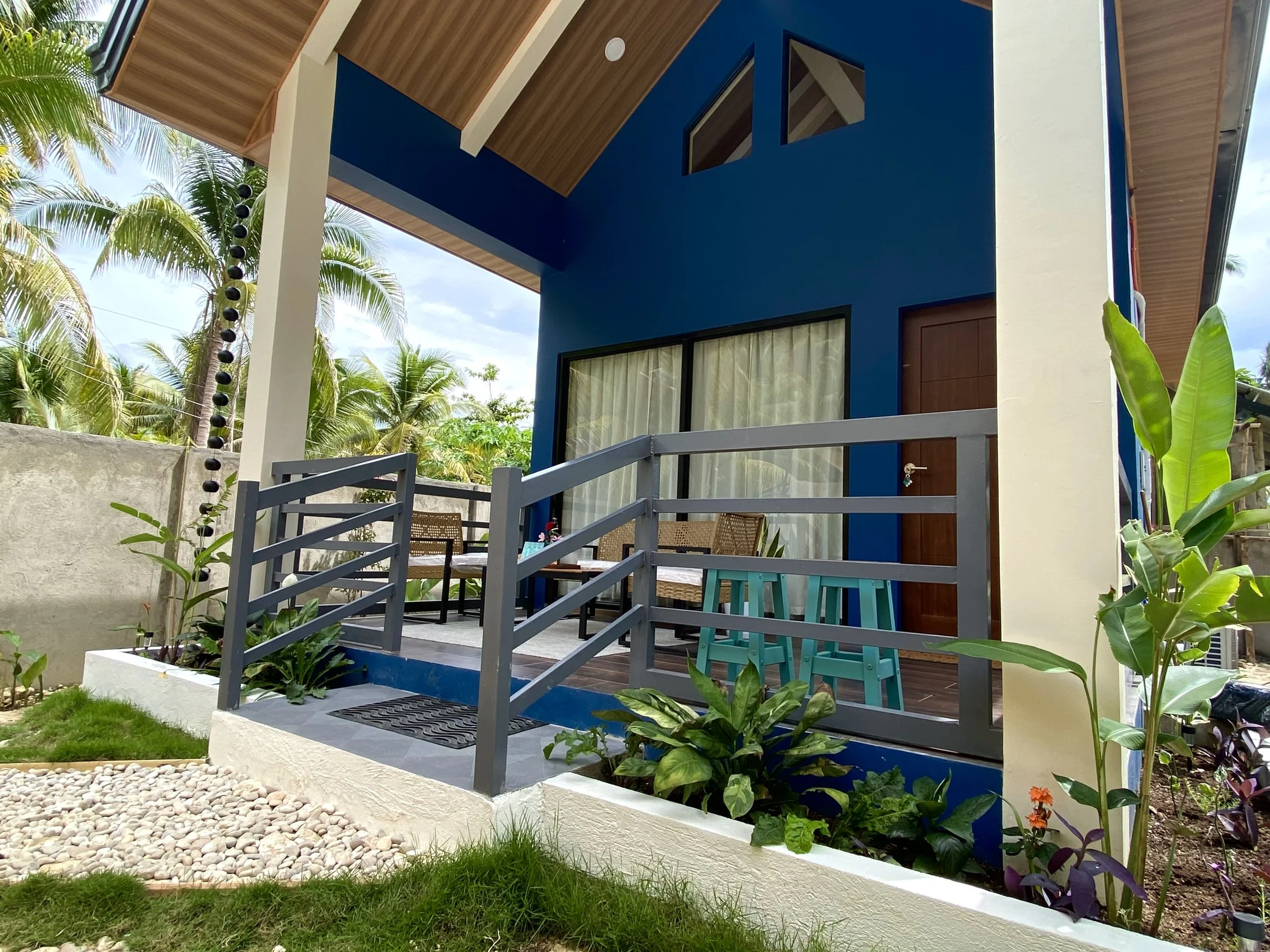 Front view of a modern house with blue walls, large windows with curtains, and a wooden roof. There is a small outdoor patio with tables and chairs, surrounded by greenery and tropical plants, including palm trees.