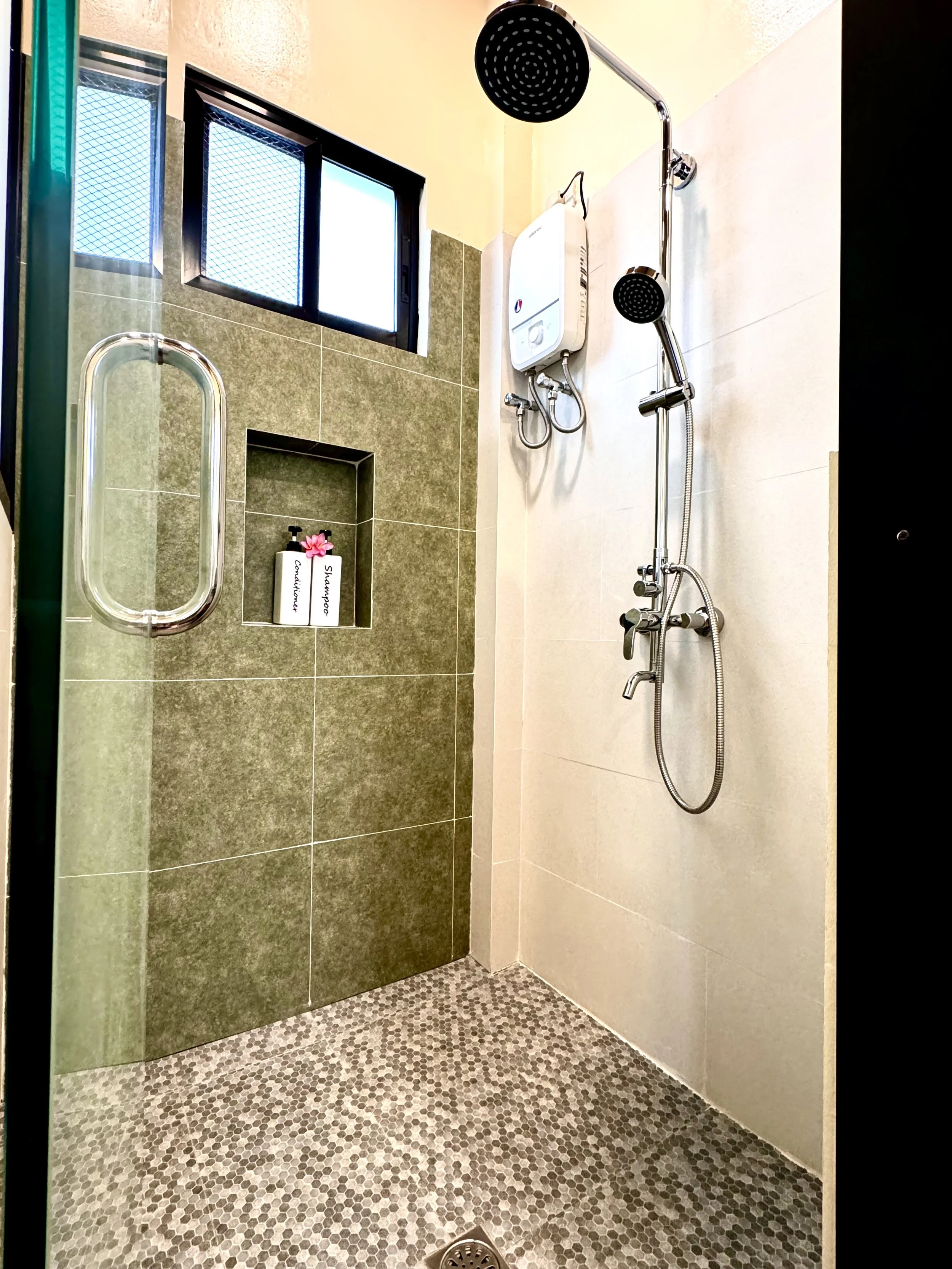 Bathroom shower with a rain shower head, handheld shower, and tiled wall with a niche containing soap and shampoo bottles, a window with mesh screen, and pebble patterned floor.