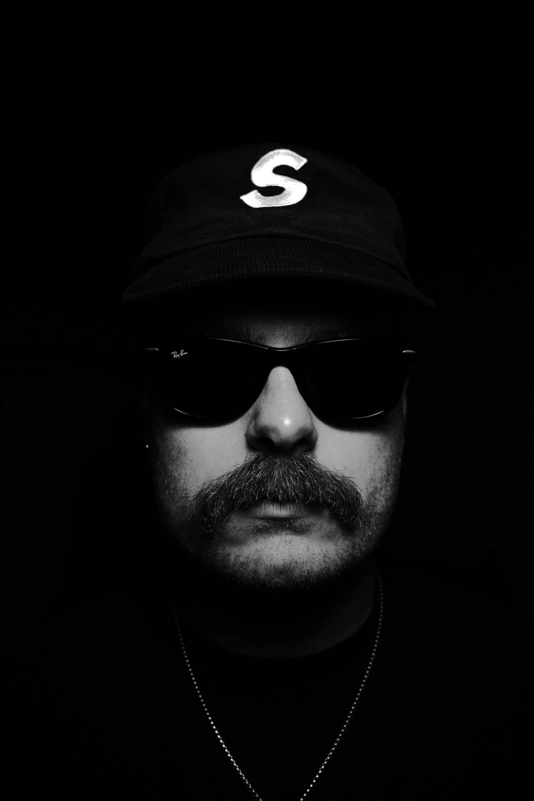 A person wearing sunglasses, a cap with a large 'S' on the front, and a necklace, photographed in black and white against a dark background.