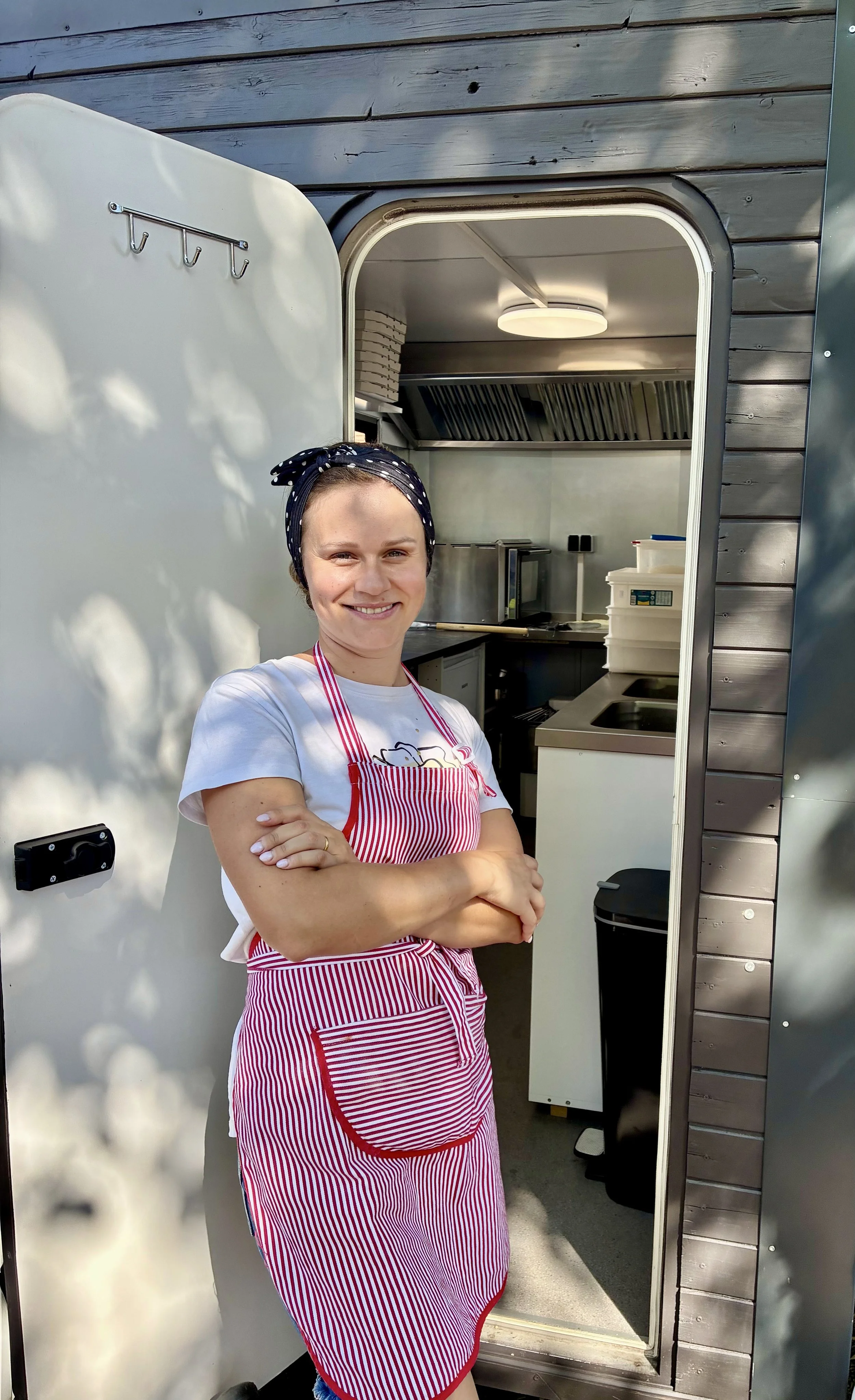 A woman wearing a black headscarf with white polka dots and a pink-striped apron is standing outdoors and smiling, with her arms crossed. She is in front of a small food truck or mobile kitchen with an open door, showing the kitchen inside.