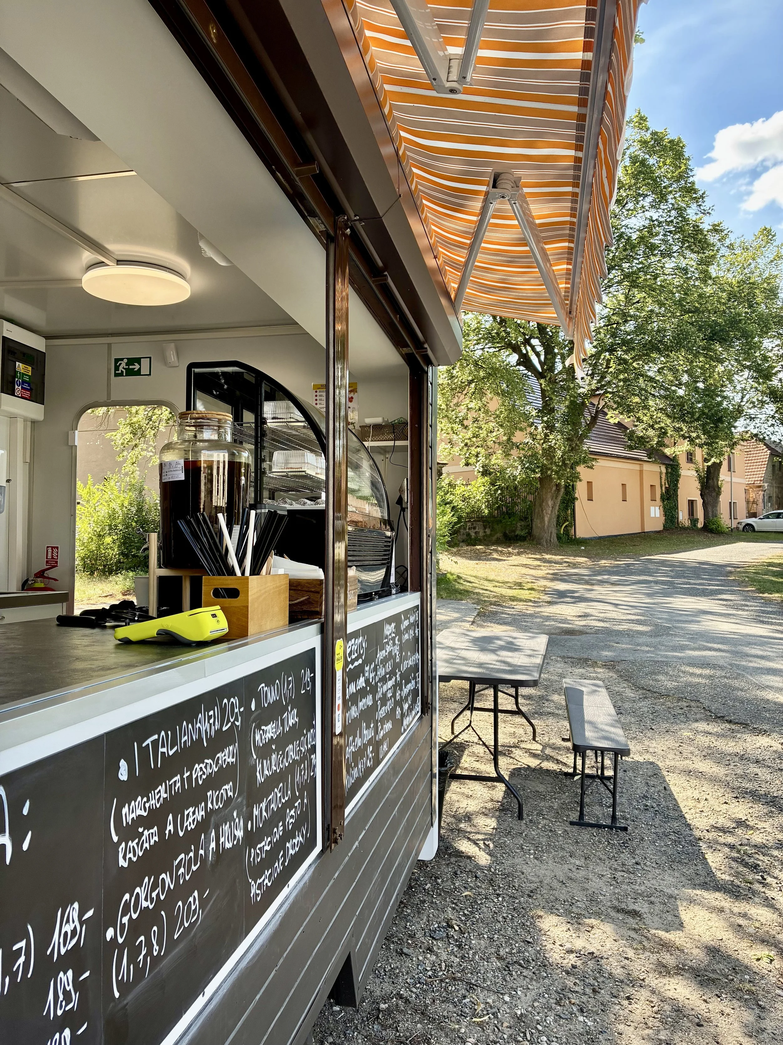 An outdoor food stand with a blackboard menu, a yellow scanner, and chairs nearby under an orange striped awning. Trees and a residential building are visible in the background on a sunny day.